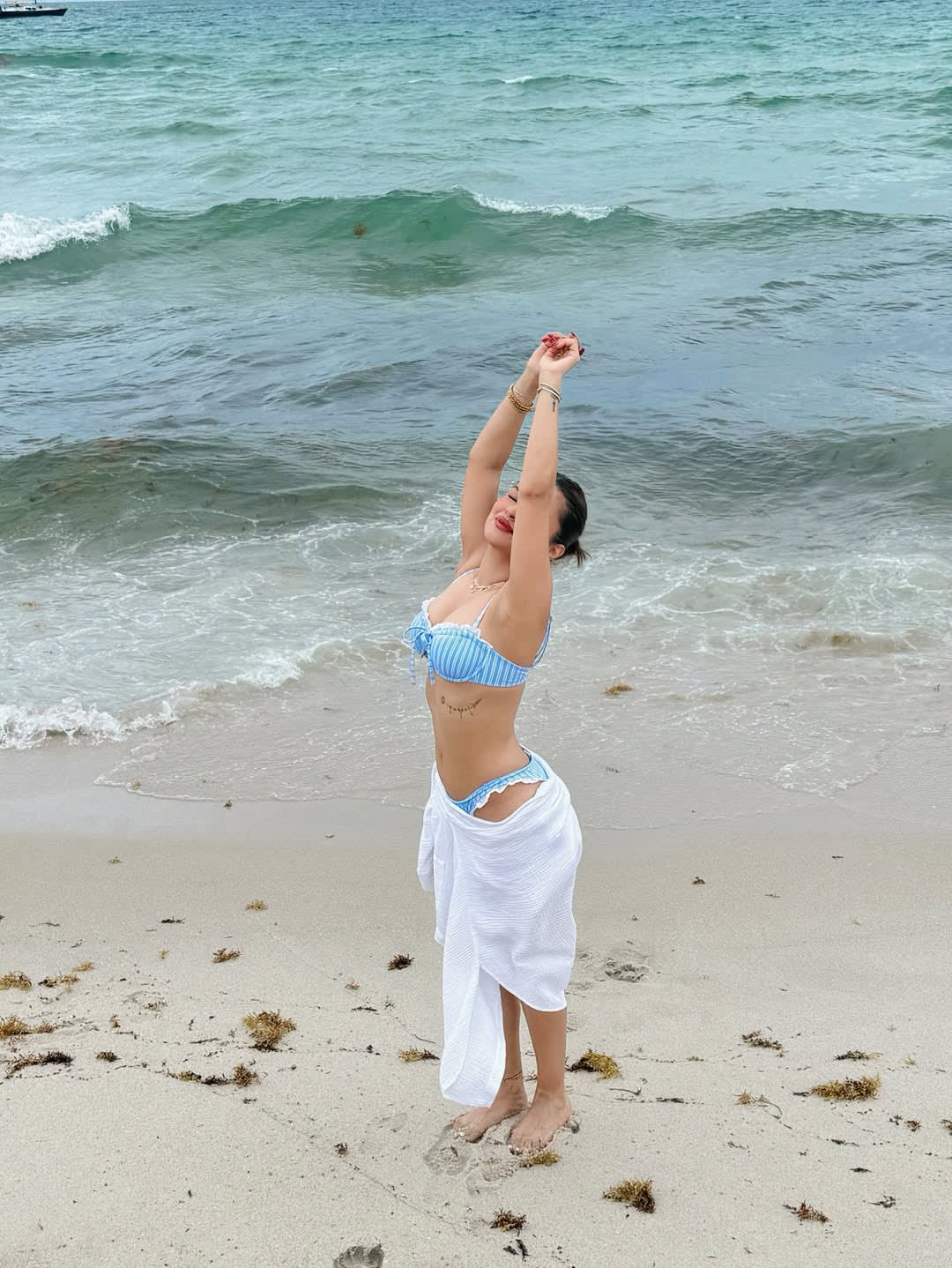 Avneet Kaur on a sandy beach, wearing a blue and white striped bikini and white sarong, stretching her arms overhead with a joyful smile, ocean waves in the background.