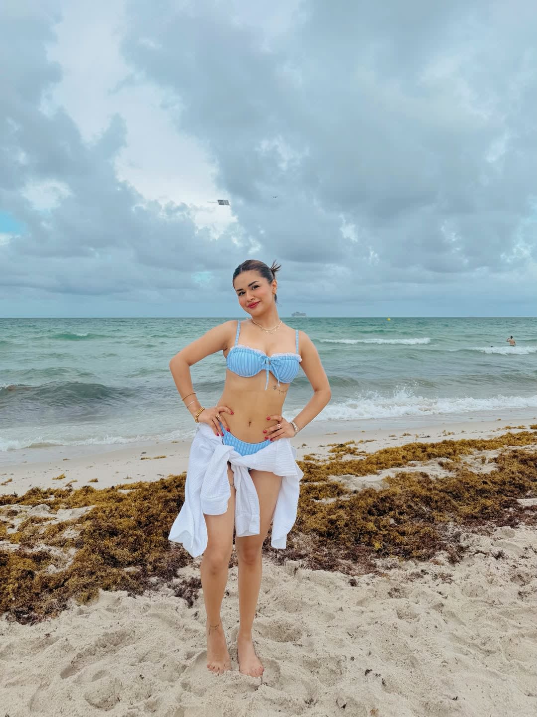Avneet Kaur standing on a beach in a light blue striped bikini with a white shirt tied around her waist, hands on hips, looking confidently at the camera. The ocean and cloudy sky are in the background.