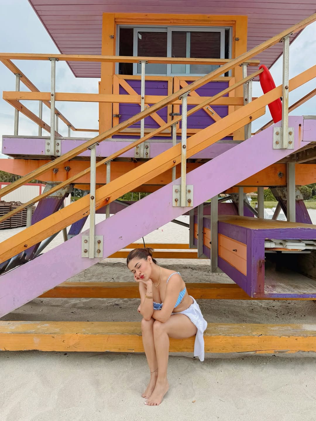 Avneet Kaur sitting on a wooden bench on a sandy beach, wearing a blue and white striped bikini top and white sarong, with a colorful orange, purple, and pink lifeguard stand in the background.