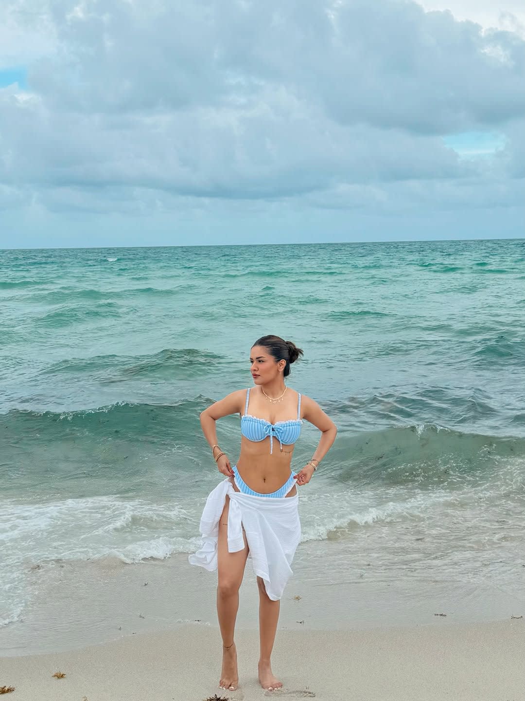 Avneet Kaur standing on a sandy beach in a light blue and white striped bikini and white sarong, looking towards the ocean under a cloudy sky.