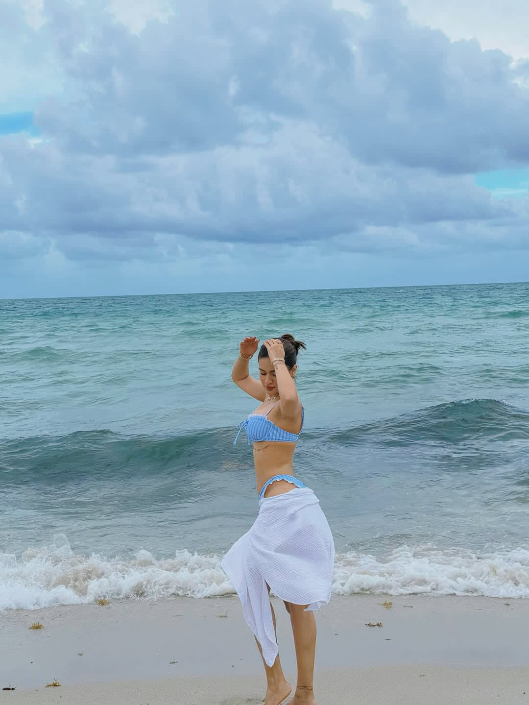 Avneet Kaur on a sandy beach, wearing a blue and white striped bikini top and a white sarong, with her hands raised, posing playfully by the ocean under a cloudy sky.