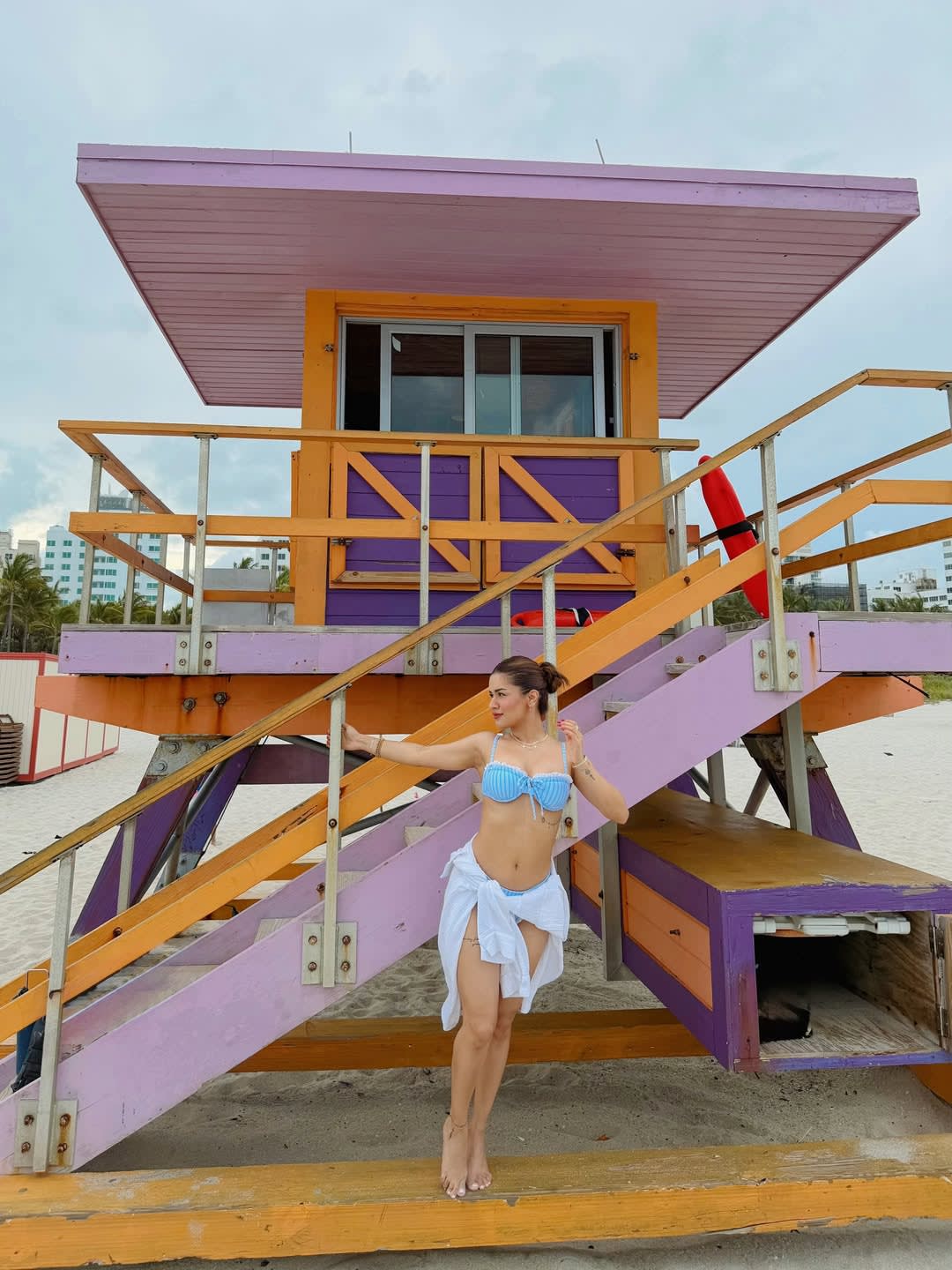Avneet Kaur in a blue and white striped bikini and white sarong, posing barefoot on a wooden step of a colorful purple, orange, and pink lifeguard stand on a sandy beach.