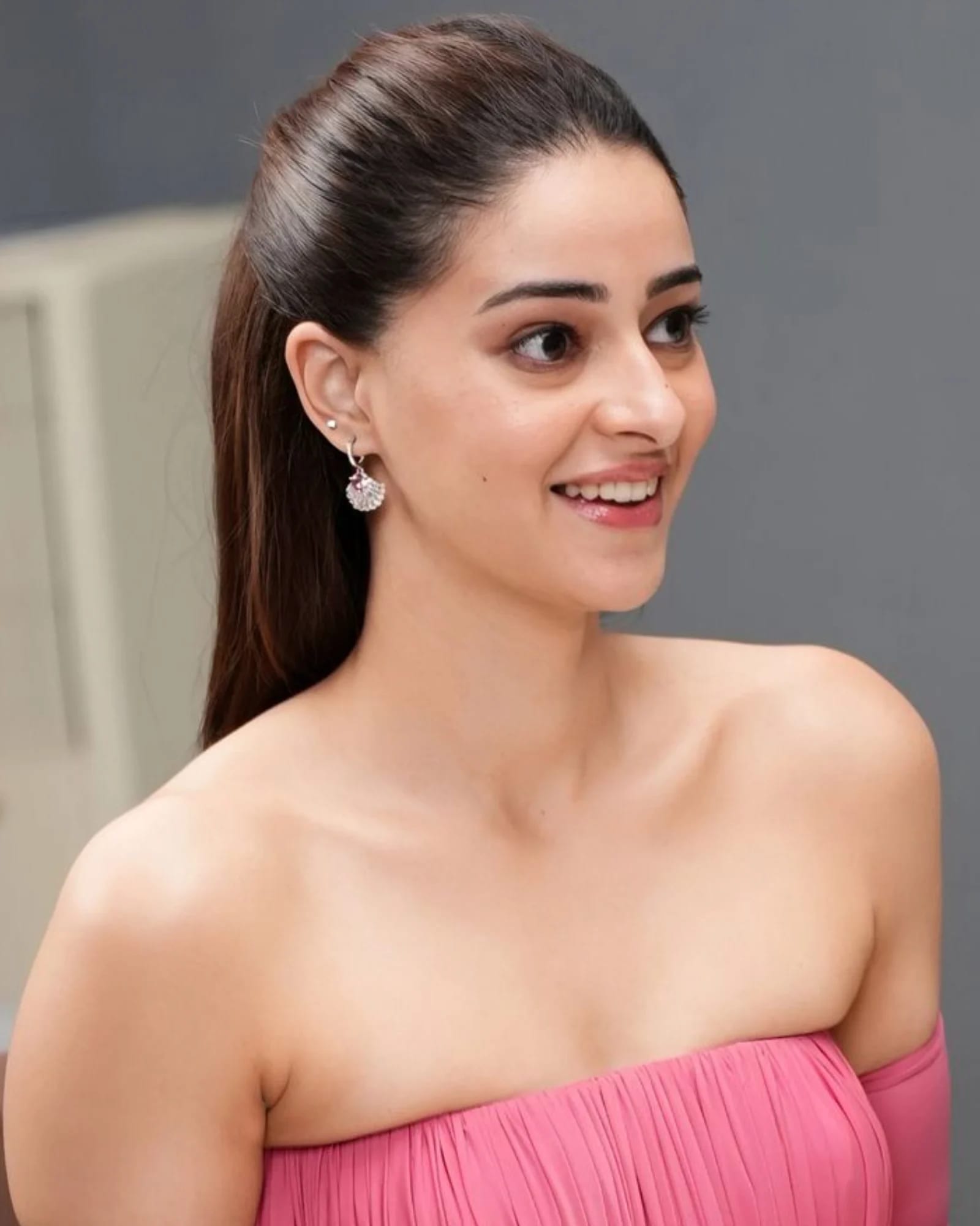 Ananya Panday smiling, wearing a pink off-shoulder pleated top, with her hair in a high ponytail and shell-shaped earrings.