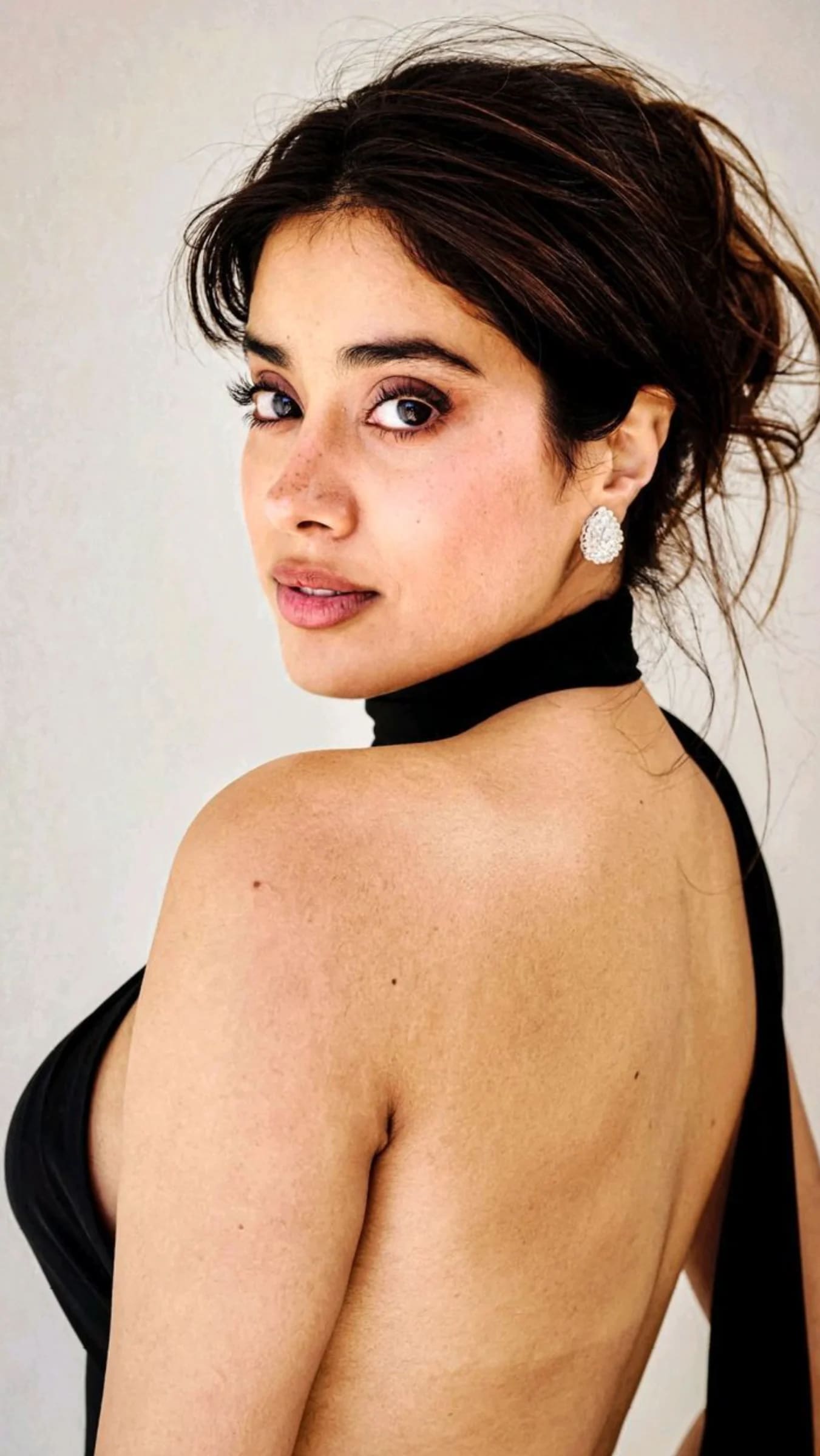 Janhvi Kapoor looking over her shoulder, wearing a black backless top and large diamond earrings, with her hair in a messy bun and visible freckles.