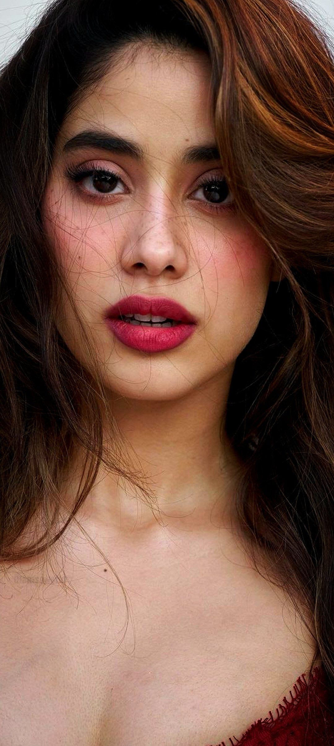 Close-up portrait of Janhvi Kapoor, an Indian actress, with long wavy brown hair, vibrant fuchsia lipstick, and expressive dark eyes, looking directly at the camera.