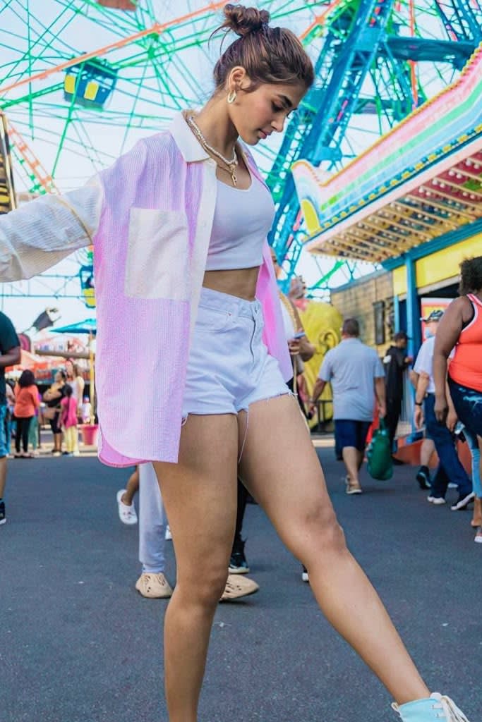Pooja Hegde, with her hair in a bun, wearing a white crop top, white denim shorts, and an open pastel pink and white striped shirt, walking at a vibrant carnival with a Ferris wheel in the background.