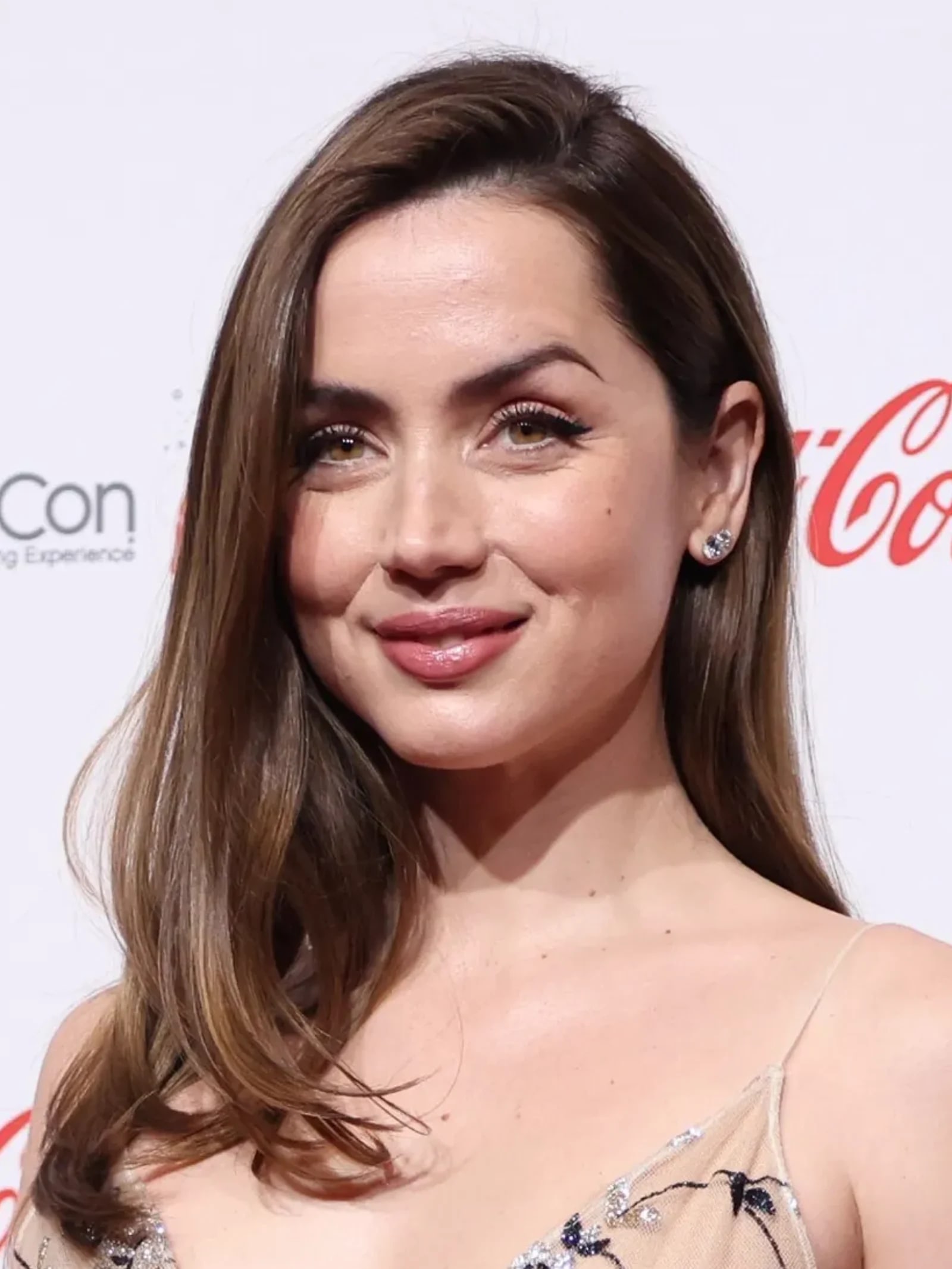 Close-up portrait of Ana de Armas smiling, wearing a nude-toned embellished dress and stud earrings at an event.