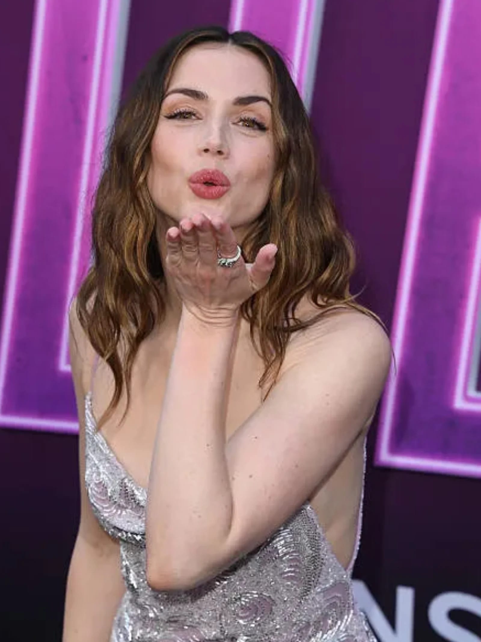 Ana de Armas, a woman with wavy brown hair, wearing a shimmering silver sequined dress, blowing a kiss towards the camera with puckered lips and a playful expression against a blurry purple background.