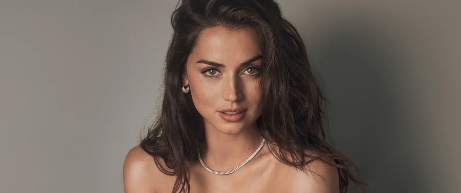 Close-up portrait of Ana de Armas with long dark hair, light brown eyes, wearing a diamond tennis necklace and small hoop earrings, against a grey background.