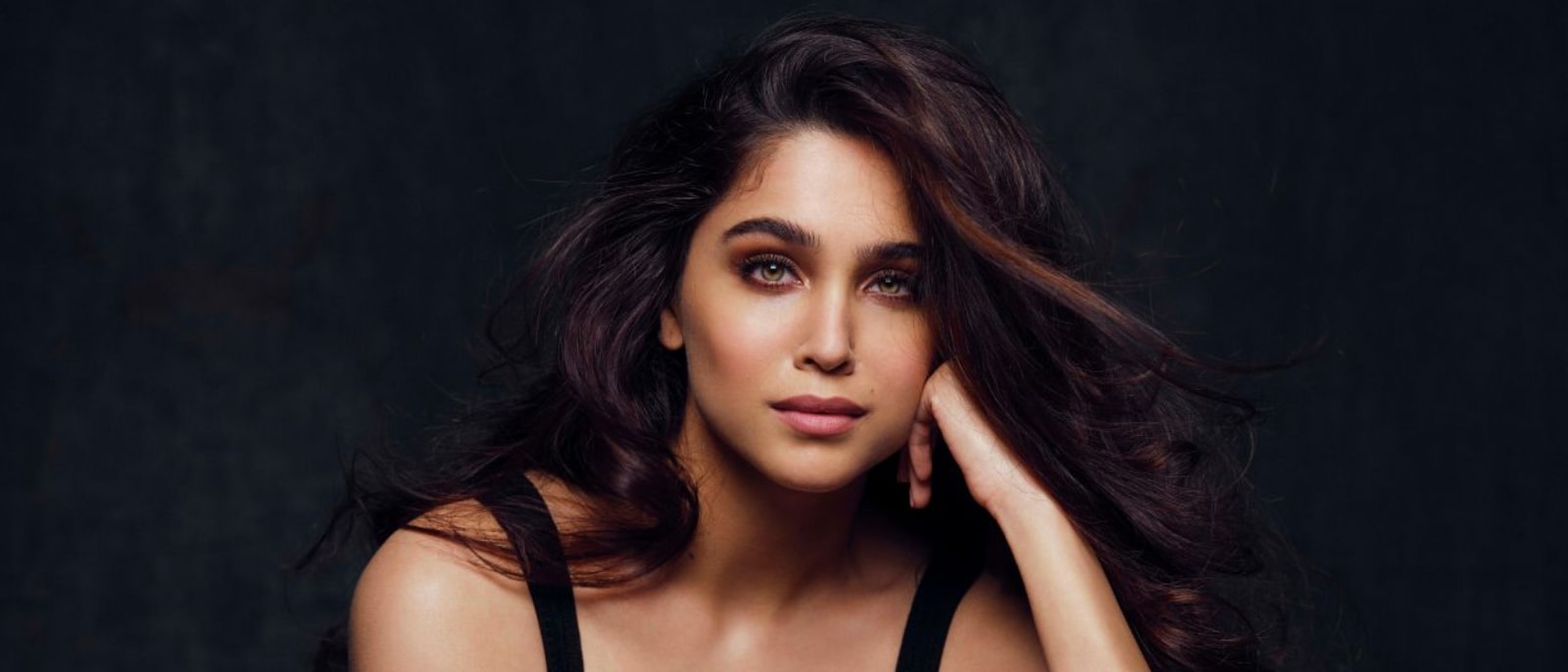 Close-up portrait of Sharvari Wagh, an Indian actress, with long dark wavy hair, light hazel eyes, and a serene expression, wearing a black top against a dark background.