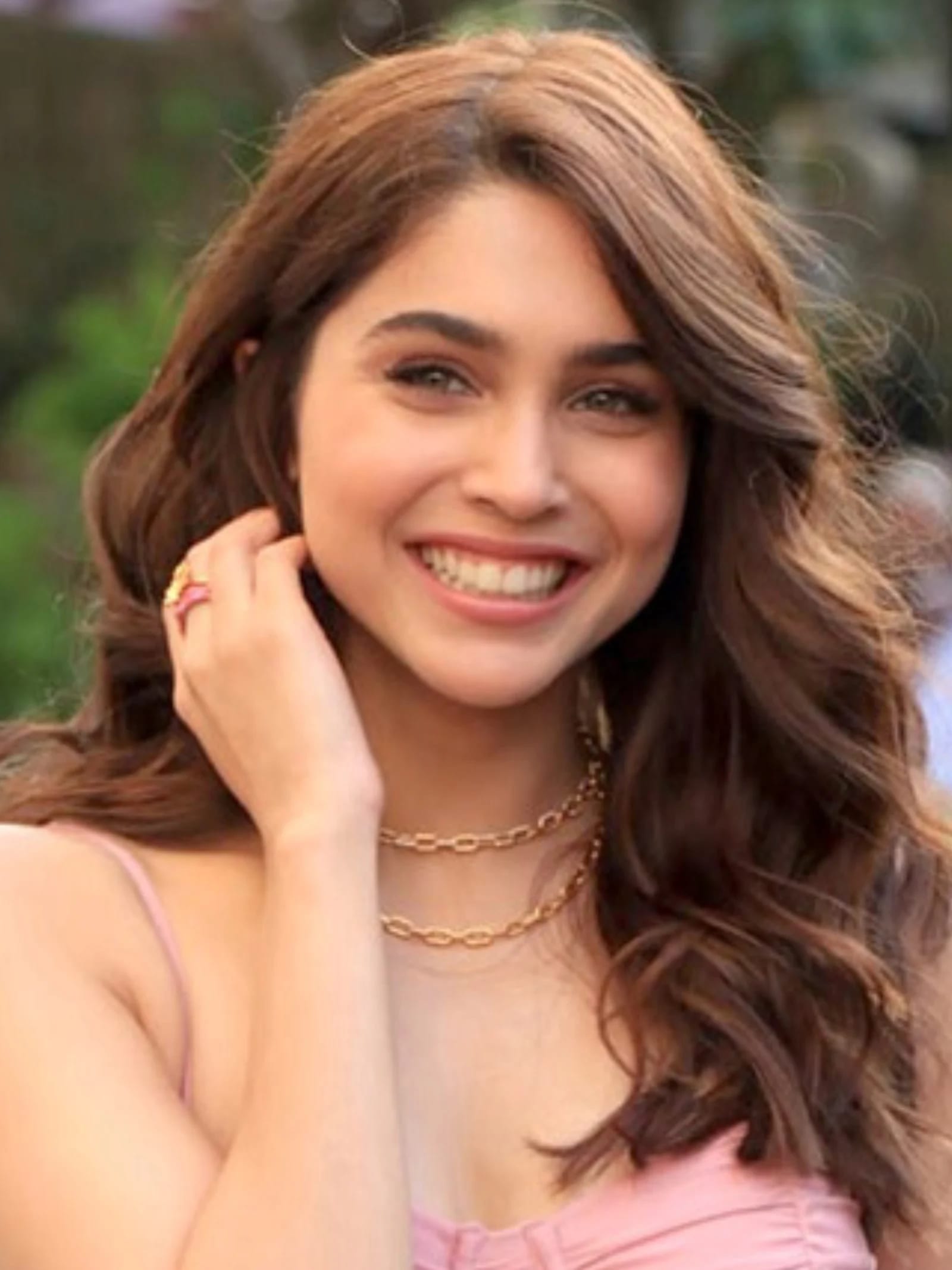Close-up portrait of Sharvari Wagh smiling brightly, wearing a pink strappy top, two gold chain necklaces, and a pink and gold ring, with wavy brown hair.
