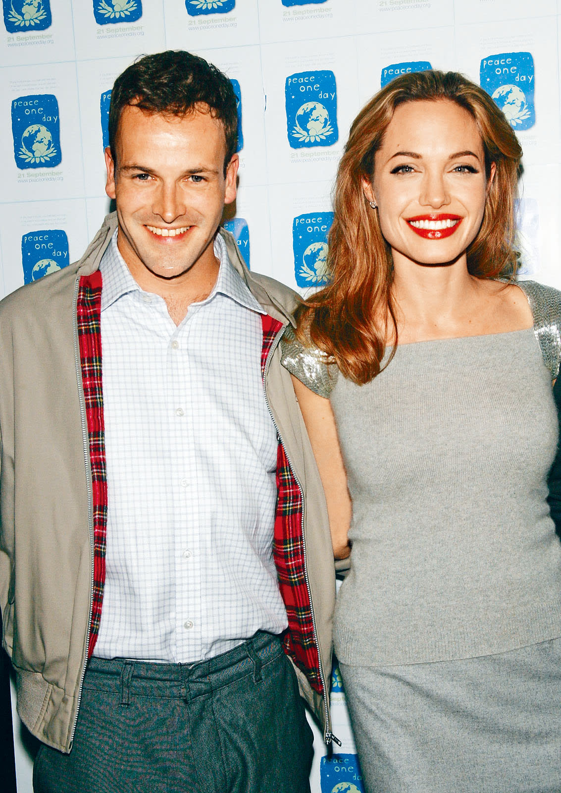 Angelina Jolie smiles alongside Jeremy Gilley at a Peace One Day event. Angelina Jolie, with long reddish-brown hair and red lipstick, smiles in a grey top with sequined sleeves next to Jeremy Gilley, who wears a plaid-lined jacket, against a 'Peace One Day' backdrop.