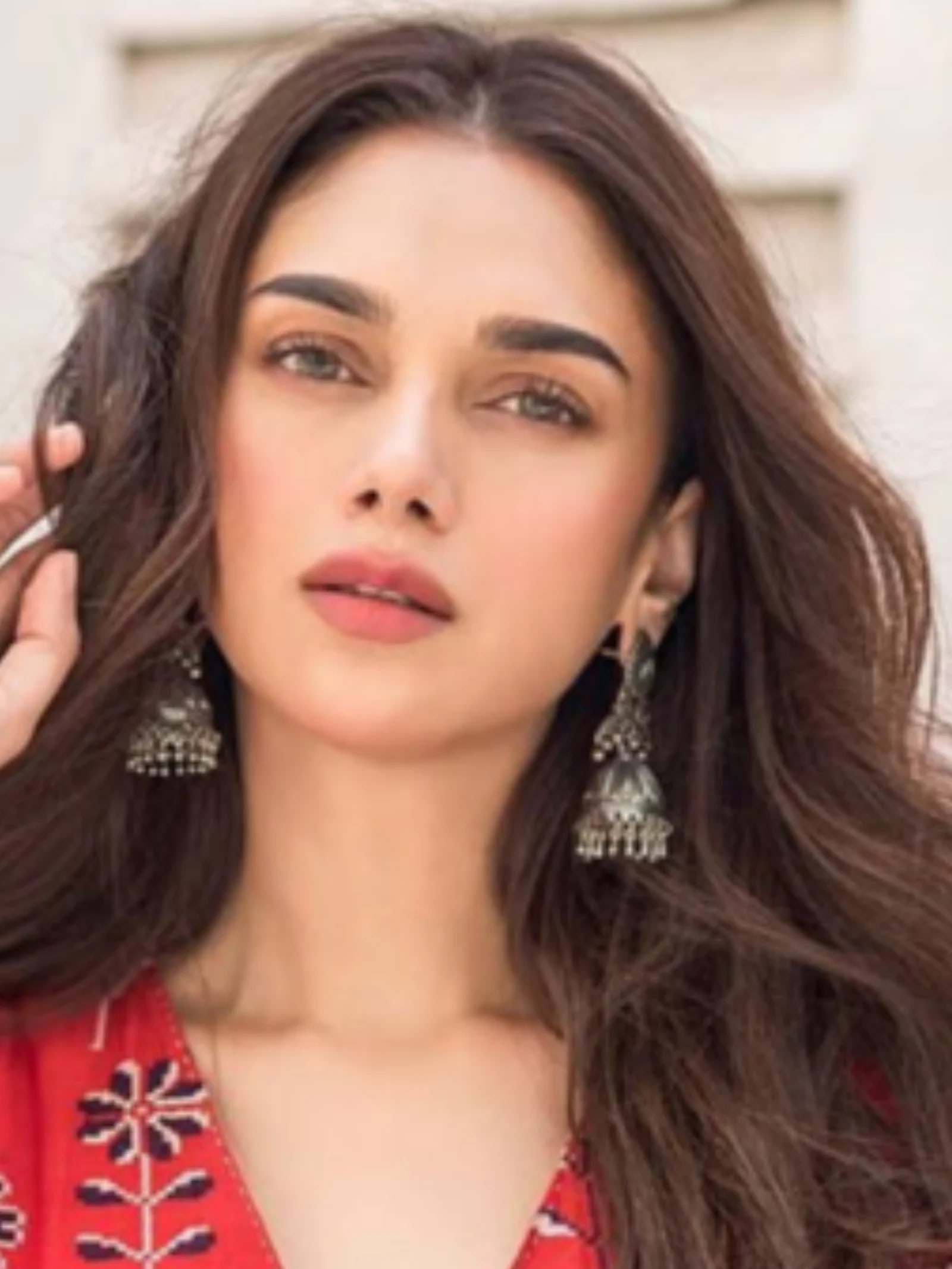 Aditi Rao Hydari in a close-up shot, wearing a red floral print outfit and silver jhumka earrings, with her hand gently touching her wavy brown hair.