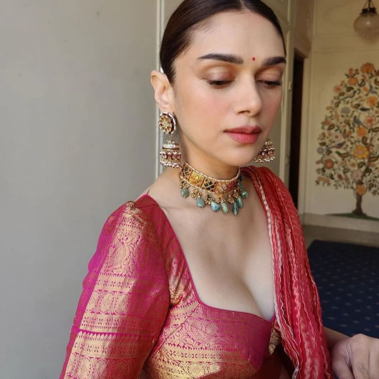 Aditi Rao Hydari in a vibrant pink and gold brocade blouse, wearing traditional jhumka earrings and a multi-colored choker necklace, with a red bindi on her forehead.
