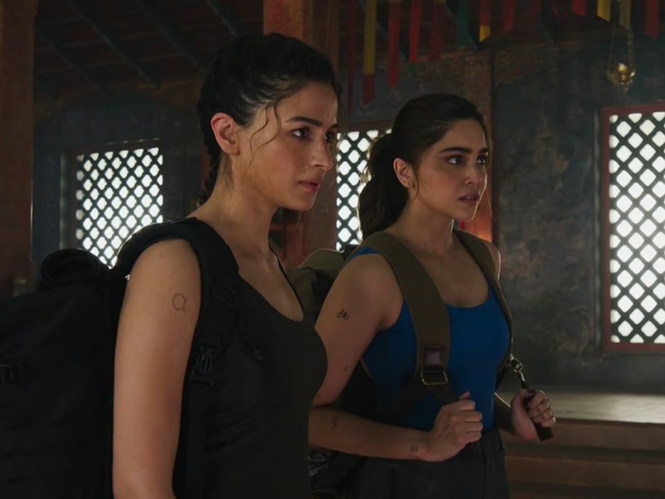 Alia Bhatt, with dark hair pulled back, wearing a dark sleeveless top and a large backpack, looking serious. An alpha tattoo is visible on her right arm.