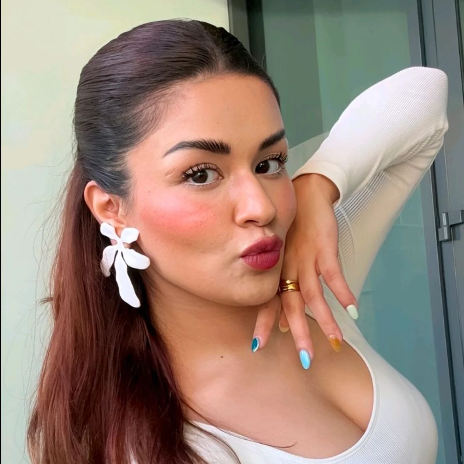 Close-up portrait of Avneet Kaur with dark brown hair, wearing a white ribbed top, large white floral earrings, and a gold ring. She has rosy blush, white eyeliner, berry lipstick, and multicolored nails, striking a playful pout.
