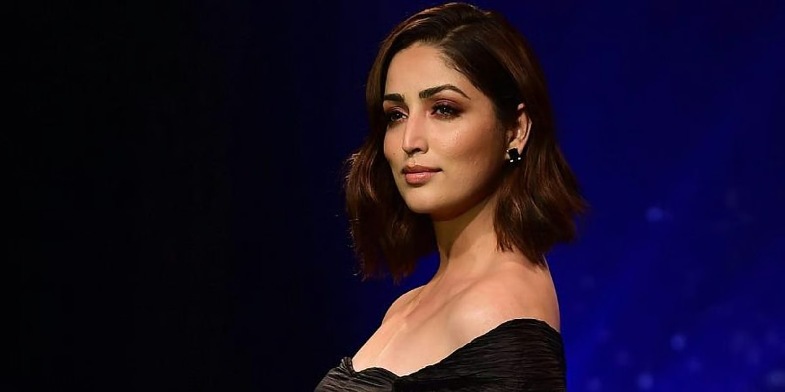 Yami Gautam posing elegantly in a black off-shoulder dress with short brown hair and sophisticated makeup against a deep blue background.