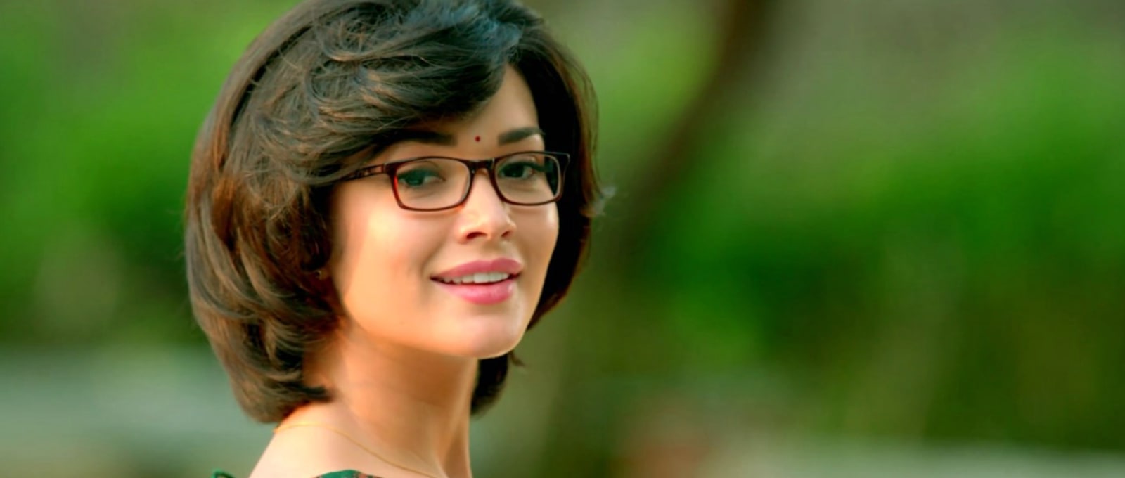 Amy Jackson, an Indian actress, smiling gently while wearing dark-framed glasses, a red bindi, and a short, wavy brown bob haircut, against a blurred vibrant green background.