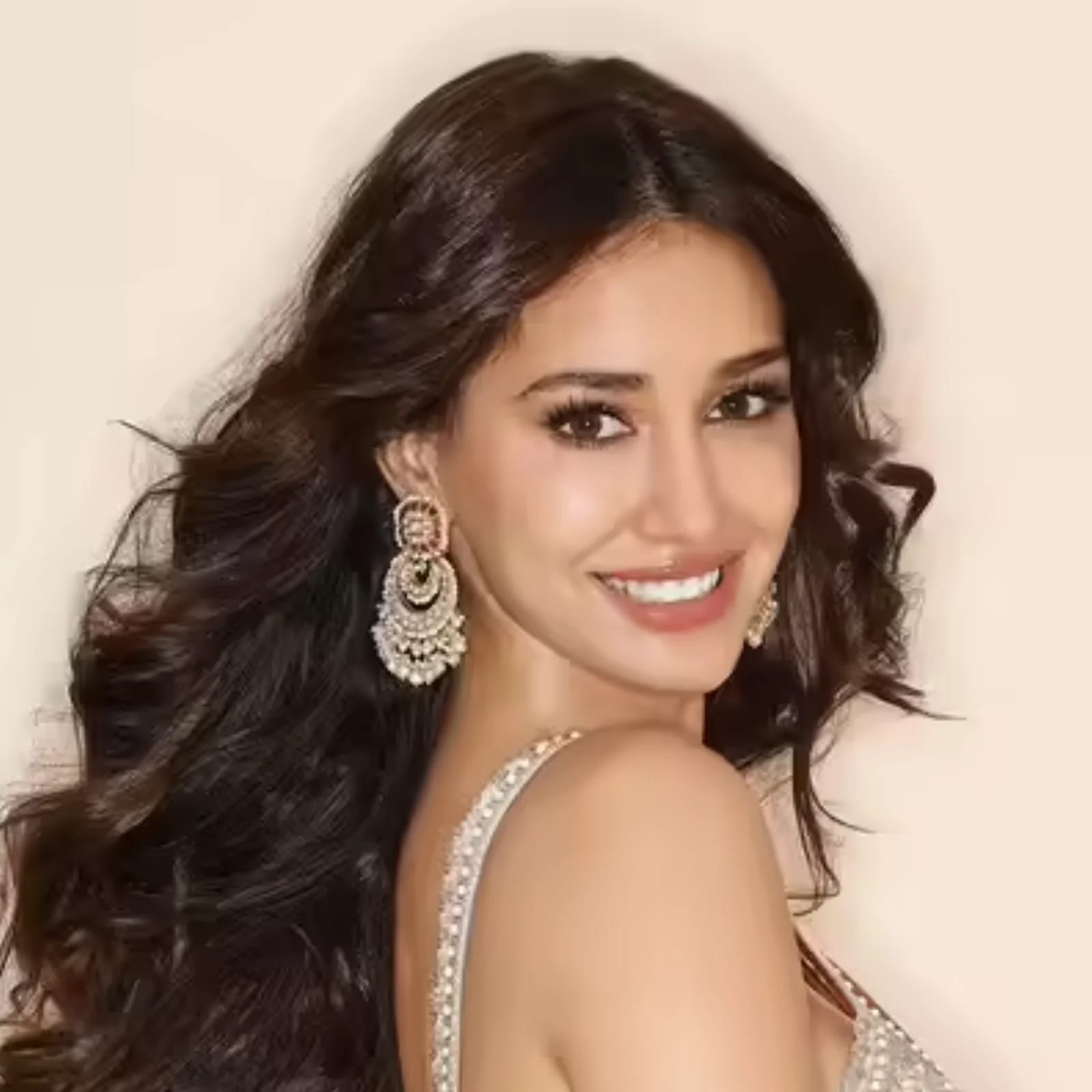 Close-up portrait of Disha Patani smiling, wearing a light-colored embellished dress and large chandelier earrings, with long wavy dark hair.