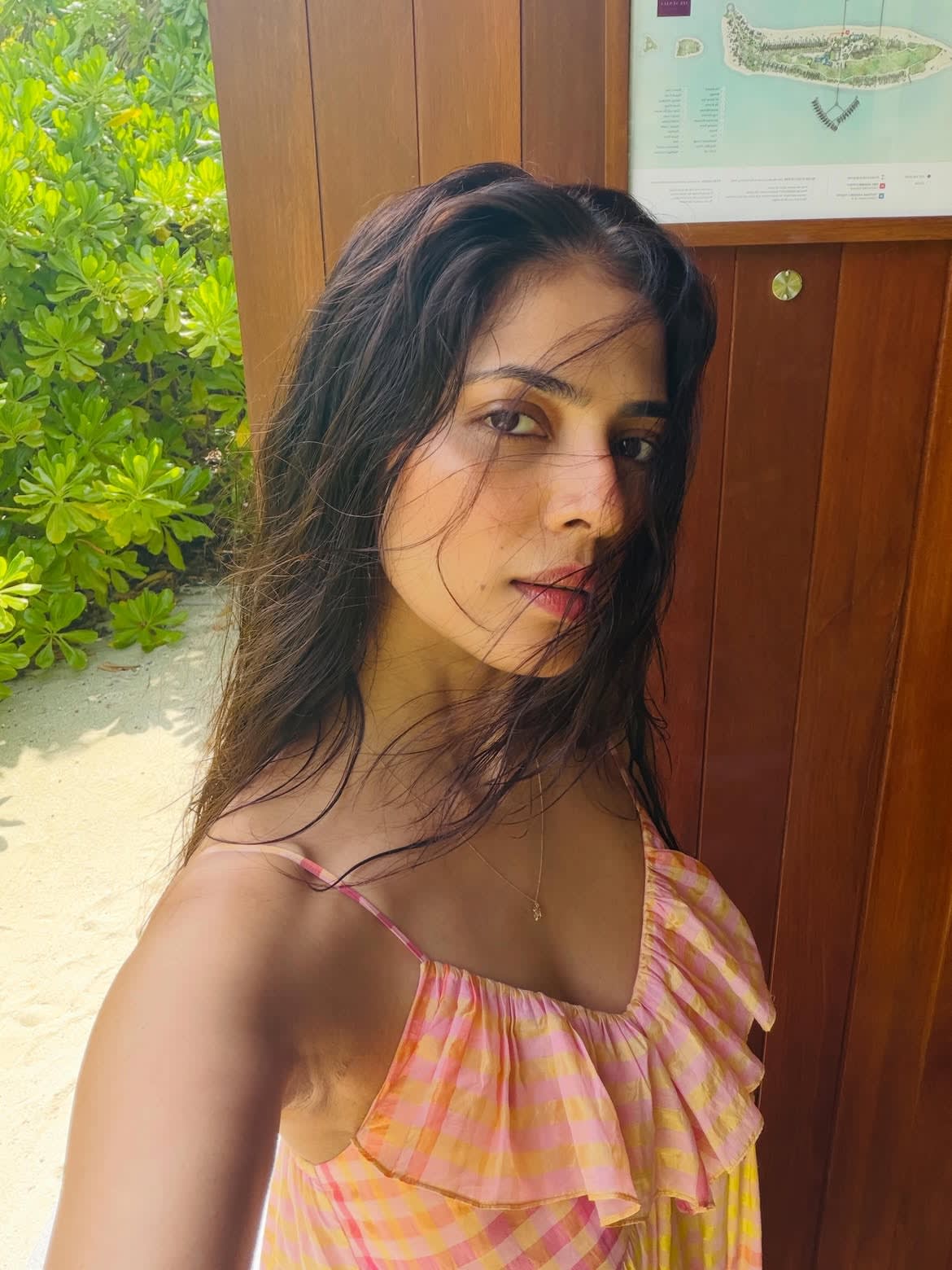 Malavika Mohanan taking a selfie in a pink and yellow plaid ruffled dress, with wet, dark hair, against a background of green tropical foliage and a wooden wall with a resort map.
