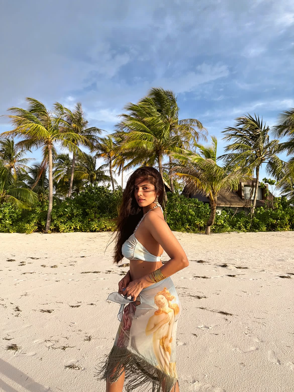Malavika Mohanan posing on a white sand beach with palm trees, wearing a light blue bikini top and a sheer sarong with classical art print, looking confidently at the camera.