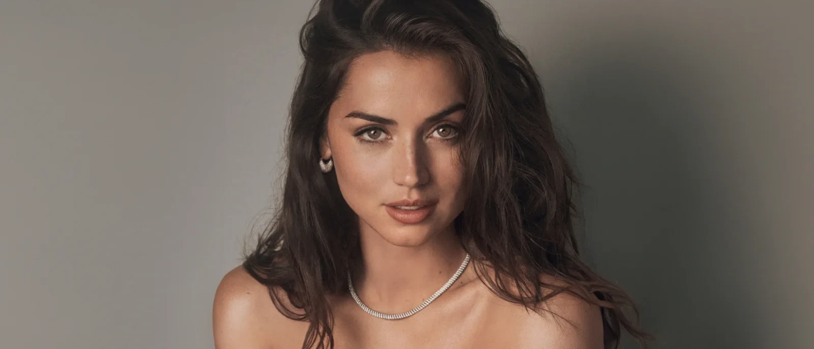 Close-up portrait of Ana de Armas with dark wavy hair, wearing a diamond necklace and small hoop earrings, looking directly at the camera with a soft expression against a grey background.