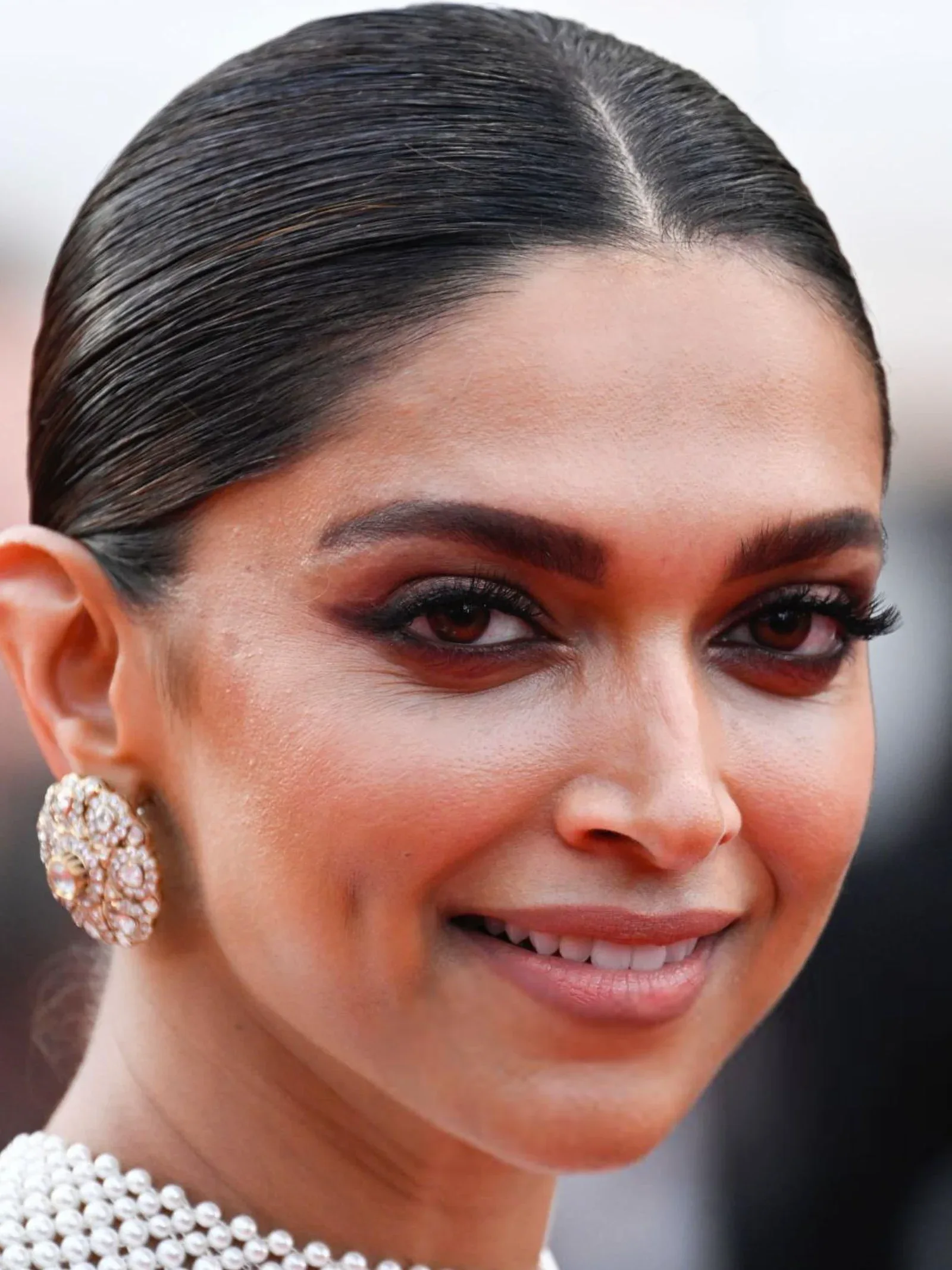 Close-up portrait of Deepika Padukone smiling, wearing dramatic winged eyeliner, large ornate gold and diamond earrings, and a multi-strand pearl necklace. Her dark hair is pulled back sleekly.