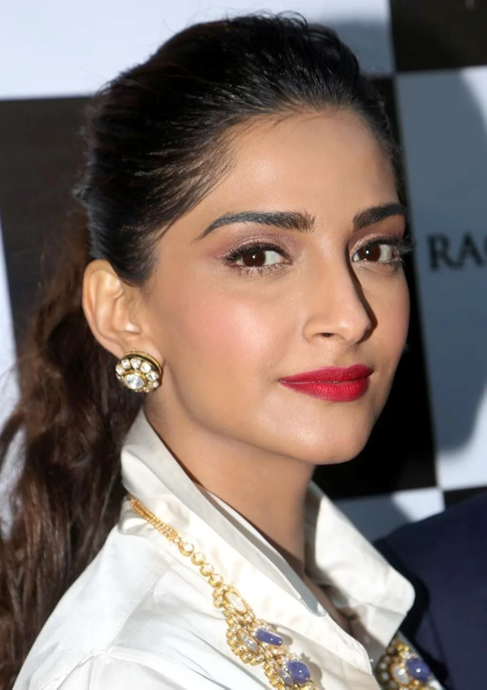 Close-up portrait of Sonam Kapoor, an Indian actress, wearing a white collared shirt, a prominent gold necklace with purple and clear stones, and large gold earrings. She has dark hair pulled back, red lipstick, and is looking directly at the camera with a confident expression.