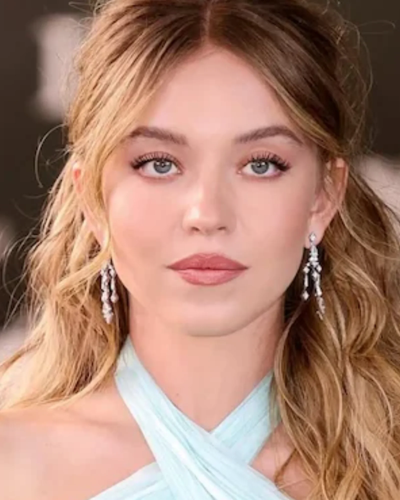 Close-up portrait of Sydney Sweeney with blonde wavy hair, striking blue eyes, wearing a mint green criss-cross top and long dangling diamond earrings, looking directly at the viewer.
