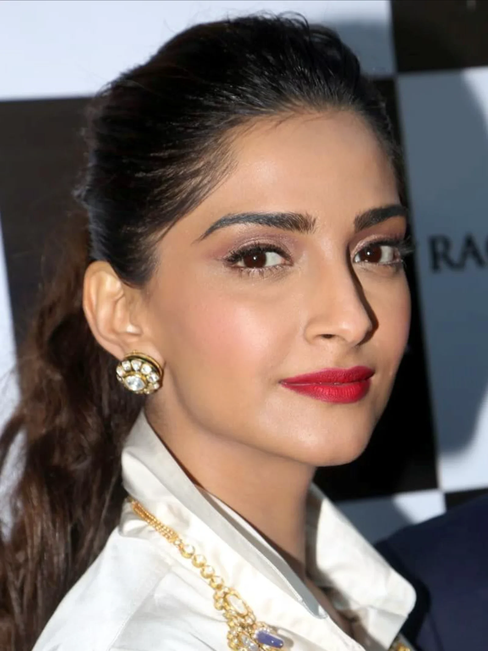 Close-up portrait of Sonam Kapoor, an Indian actress, wearing a white collared shirt, gold and diamond earrings, and a gold necklace with a purple stone. She has her dark hair pulled back, defined eyebrows, and bright red lipstick, looking directly at the camera with a slight smile.