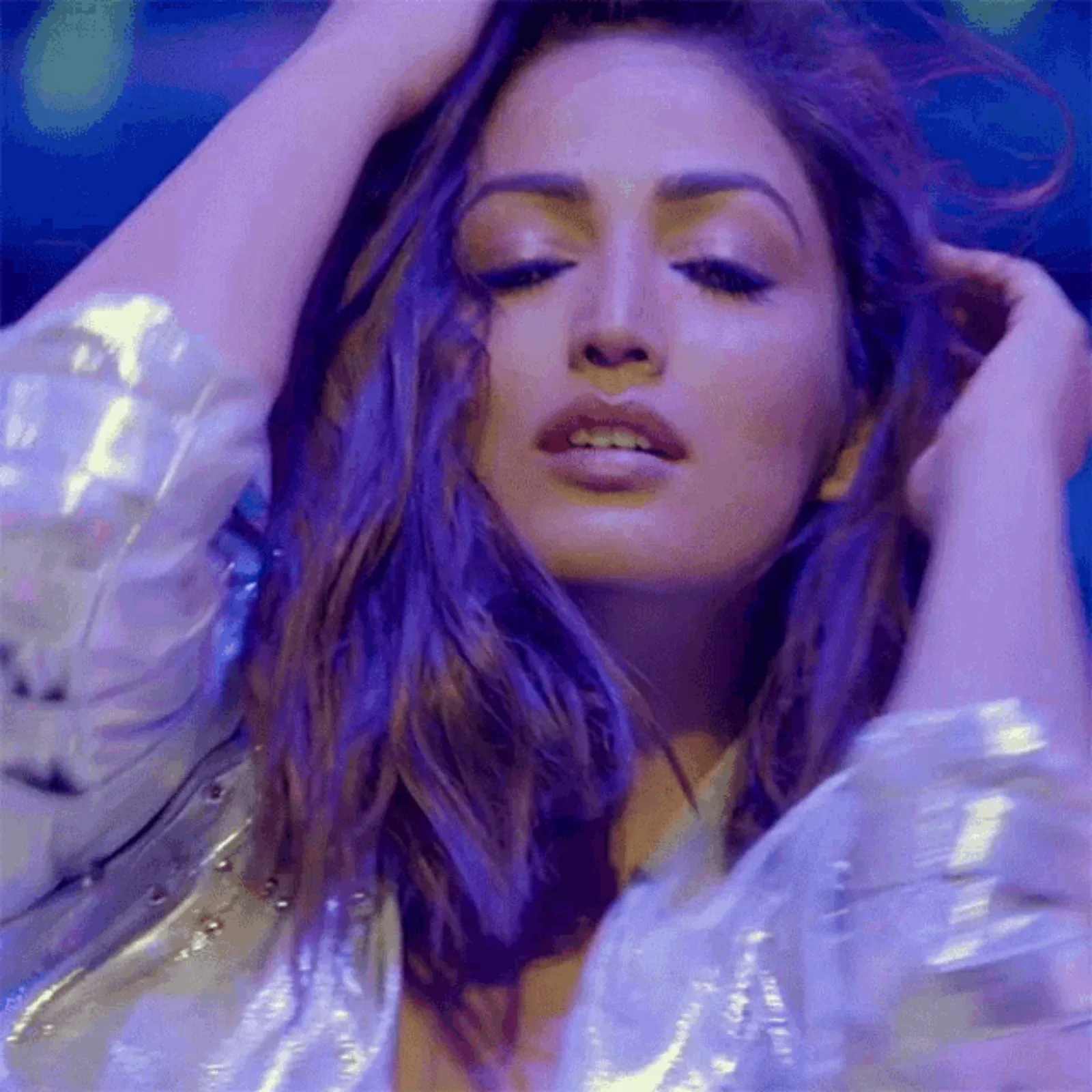 Yami Gautam with eyes closed, hands in her long wavy hair, wearing a shimmering silver jacket under blue and purple stage lighting.