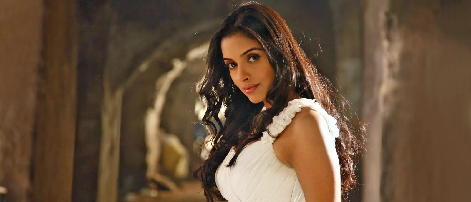 Asin Thottumkal, an Indian actress, smiling gently in a white ruffled sleeveless top with long wavy dark hair, against a warm, blurred background.