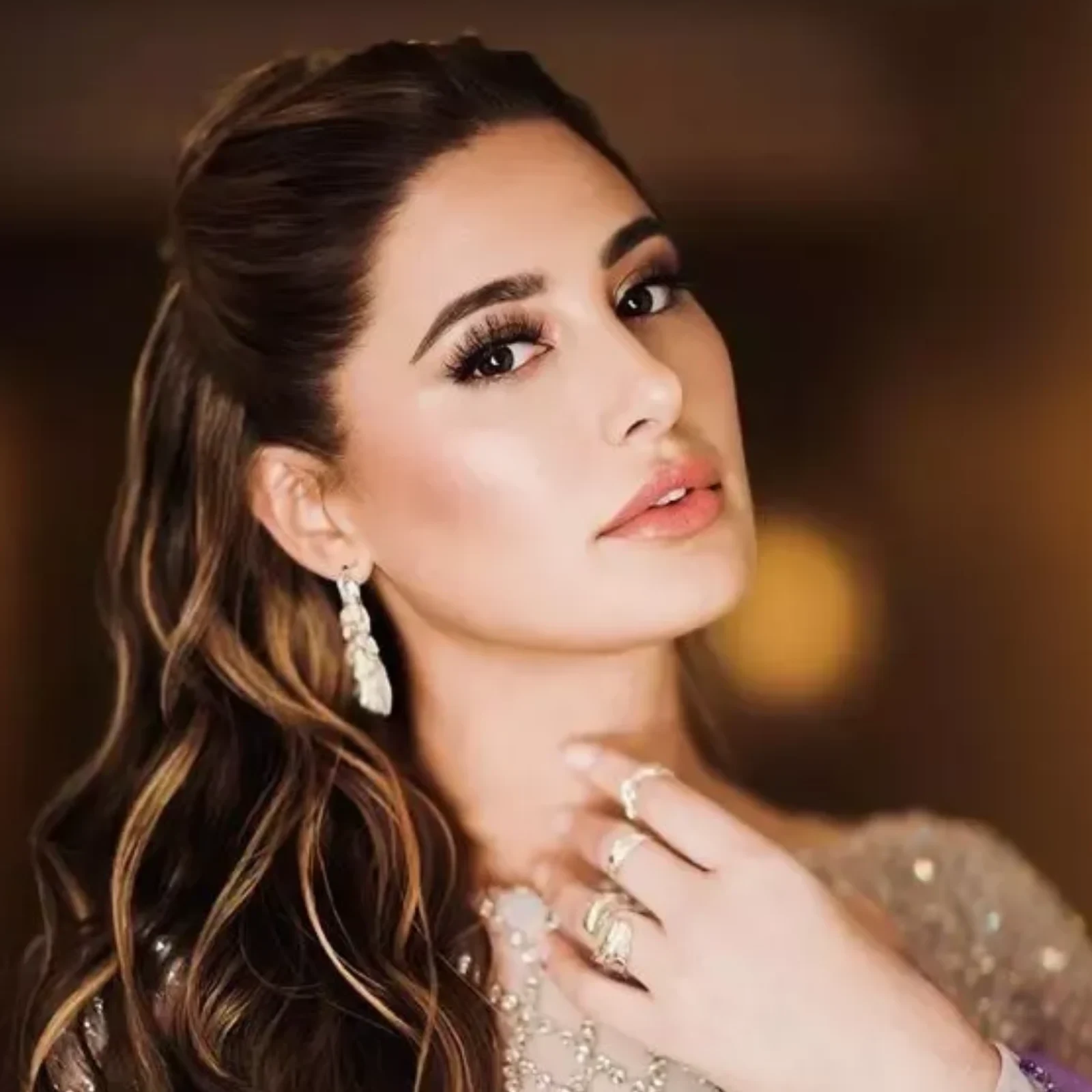 Close-up portrait of Nargis Fakhri, an Indian-American actress, with long wavy brown hair, dramatic eye makeup, pink lipstick, wearing sparkling drop earrings and rings, in an embellished outfit.