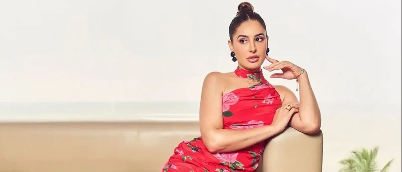 Nargis Fakhri in a red floral one-shoulder dress with a high bun, posing thoughtfully on a light sofa.