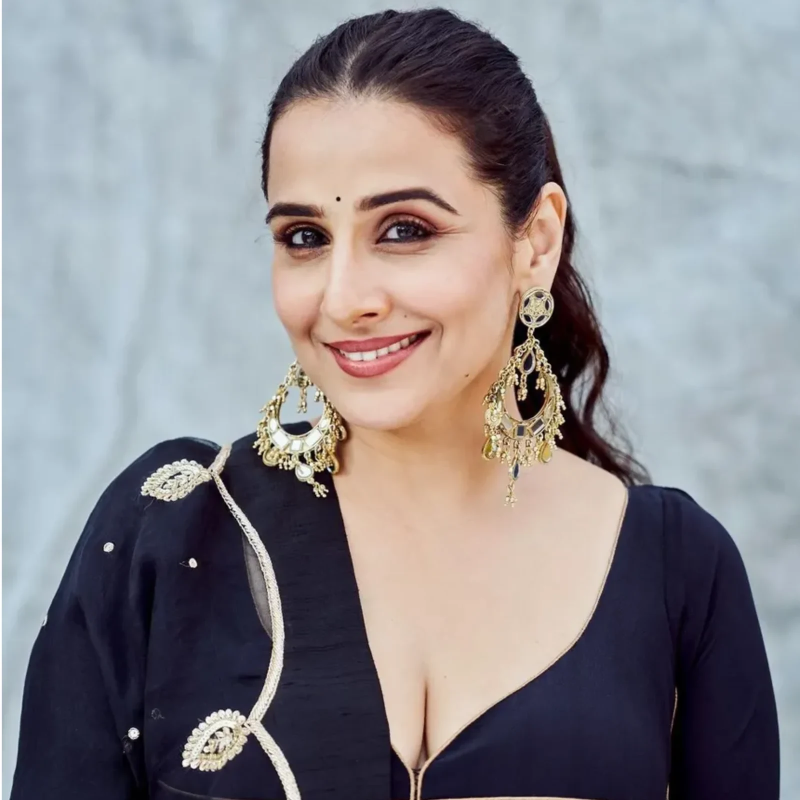 Close-up portrait of Vidya Balan smiling, wearing a black traditional outfit with gold embroidery and large gold chandelier earrings.