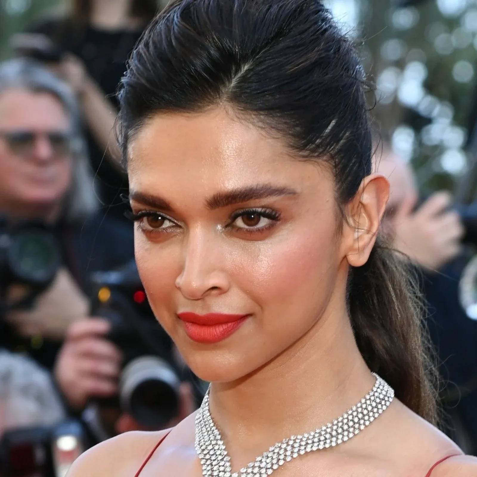 Deepika Padukone in a red dress and multi-layered diamond necklace, with her dark hair pulled back in a sleek ponytail, posing on a red carpet.