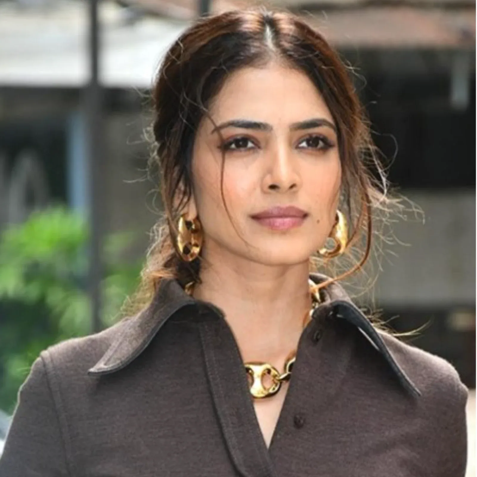 Malavika Mohanan in a close-up shot, wearing a dark brown collared shirt, large gold hoop earrings, and a chunky gold necklace, with her brown hair styled loosely.