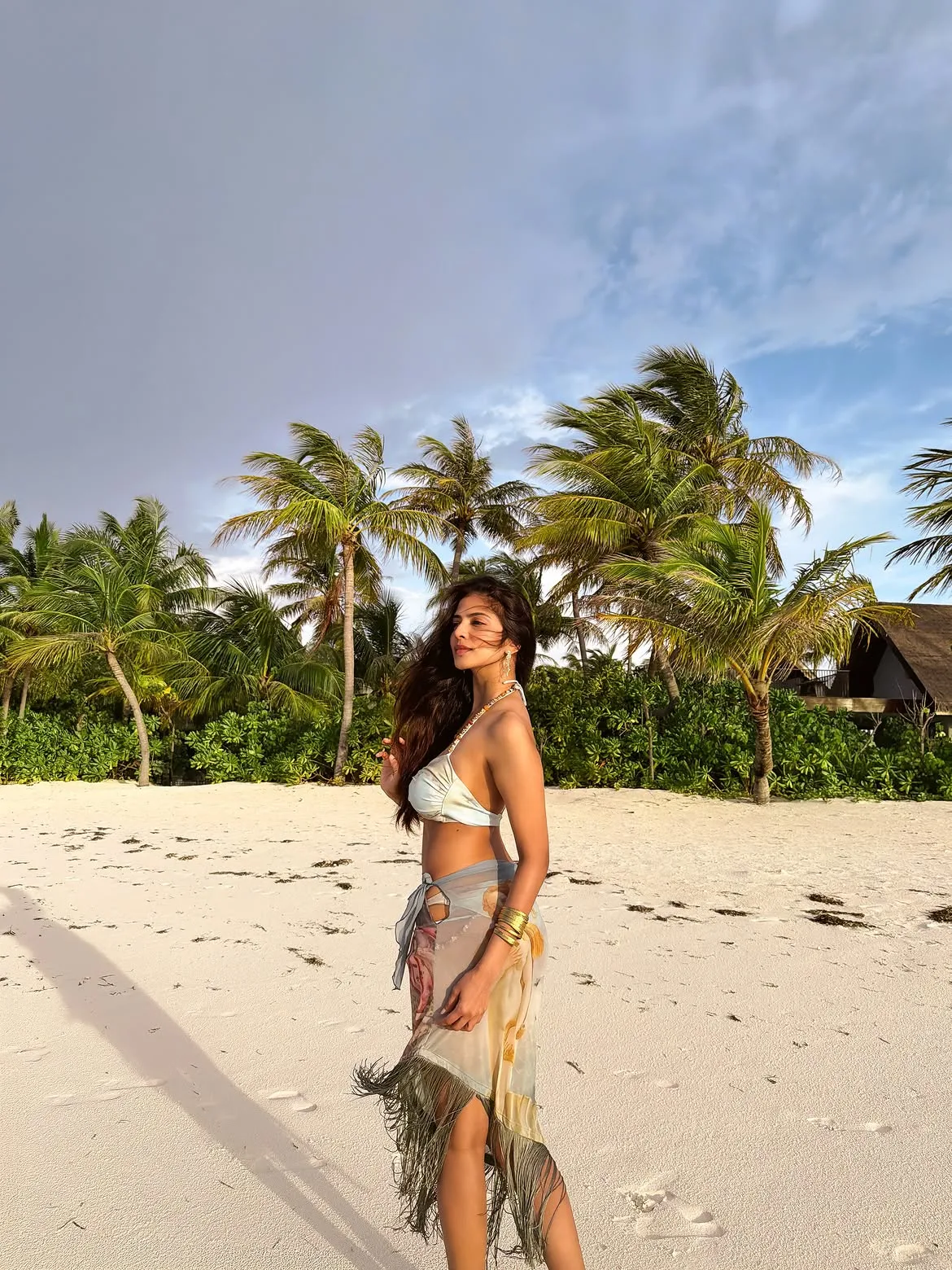 Malavika Mohanan on a white sand beach, wearing a light blue bikini top and a patterned sarong with fringe, surrounded by palm trees under a partly cloudy sky.