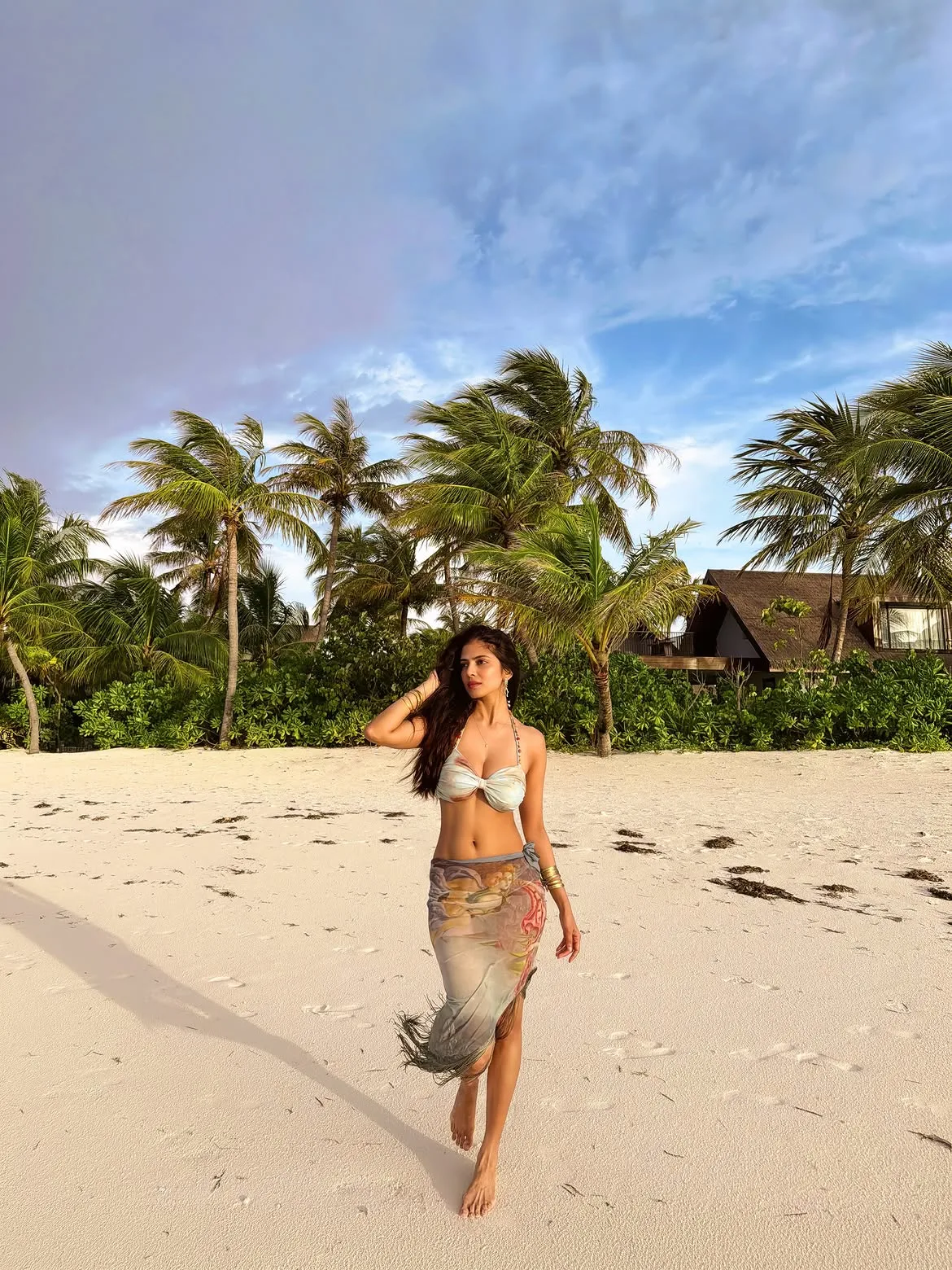 Malavika Mohanan walking barefoot on a white sand beach, wearing a light bikini top and a printed sarong, with palm trees and a resort in the background under a blue sky.
