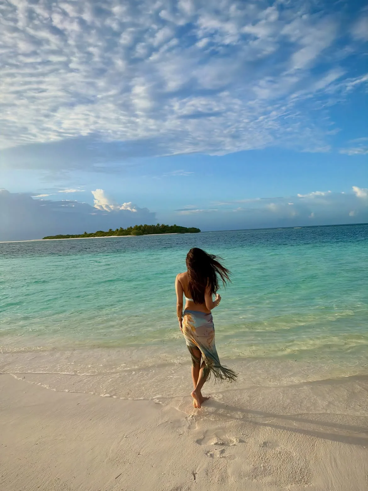 Malavika Mohanan with long dark hair, wearing a bikini top and patterned sarong, walks barefoot into the clear turquoise ocean water on a white sandy beach, with a tropical island in the distance under a blue sky.