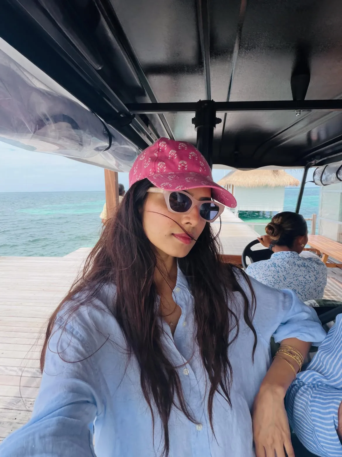 Malavika Mohanan wearing a pink patterned baseball cap, white cat-eye sunglasses, and a light blue linen shirt, sitting in a vehicle on a wooden pier over clear turquoise water in the Maldives.