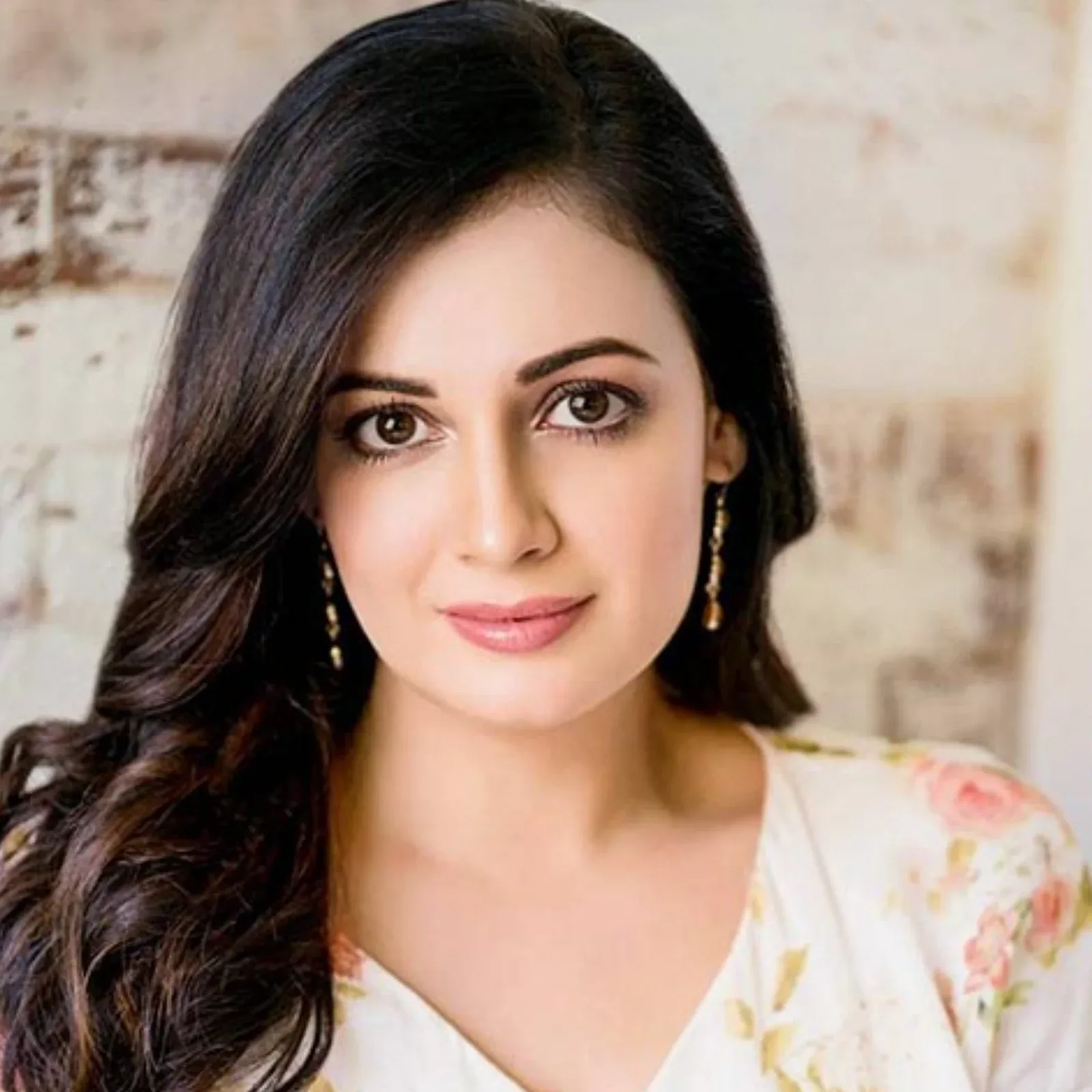 Close-up portrait of Dia Mirza, an Indian actress, smiling gently while wearing a white floral top with pink and green patterns, and gold dangle earrings. Her dark hair is styled in soft waves.