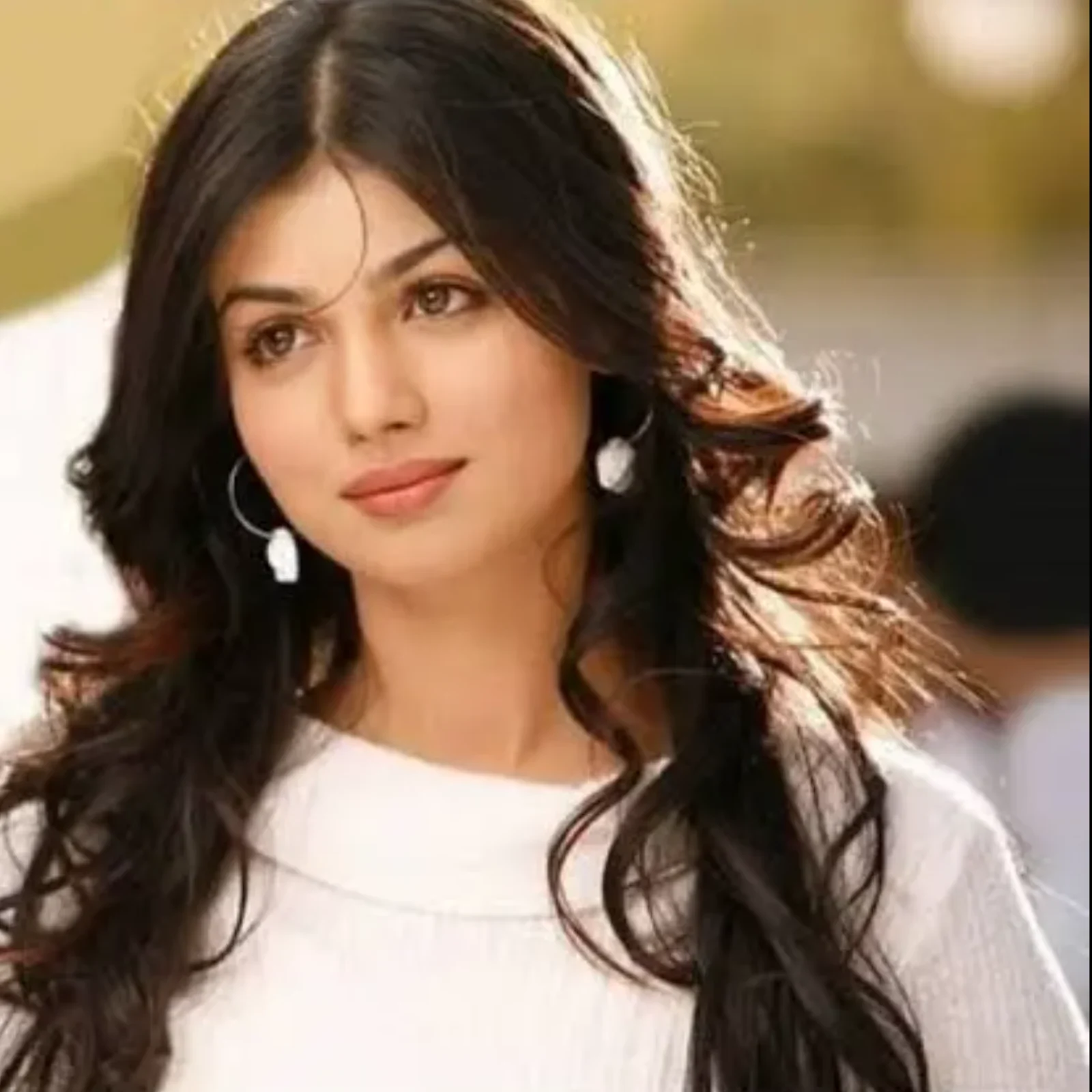 Ayesha Takia, an Indian actress, in a close-up portrait, wearing a white top, with long dark wavy hair and a gentle smile, looking slightly to the side.