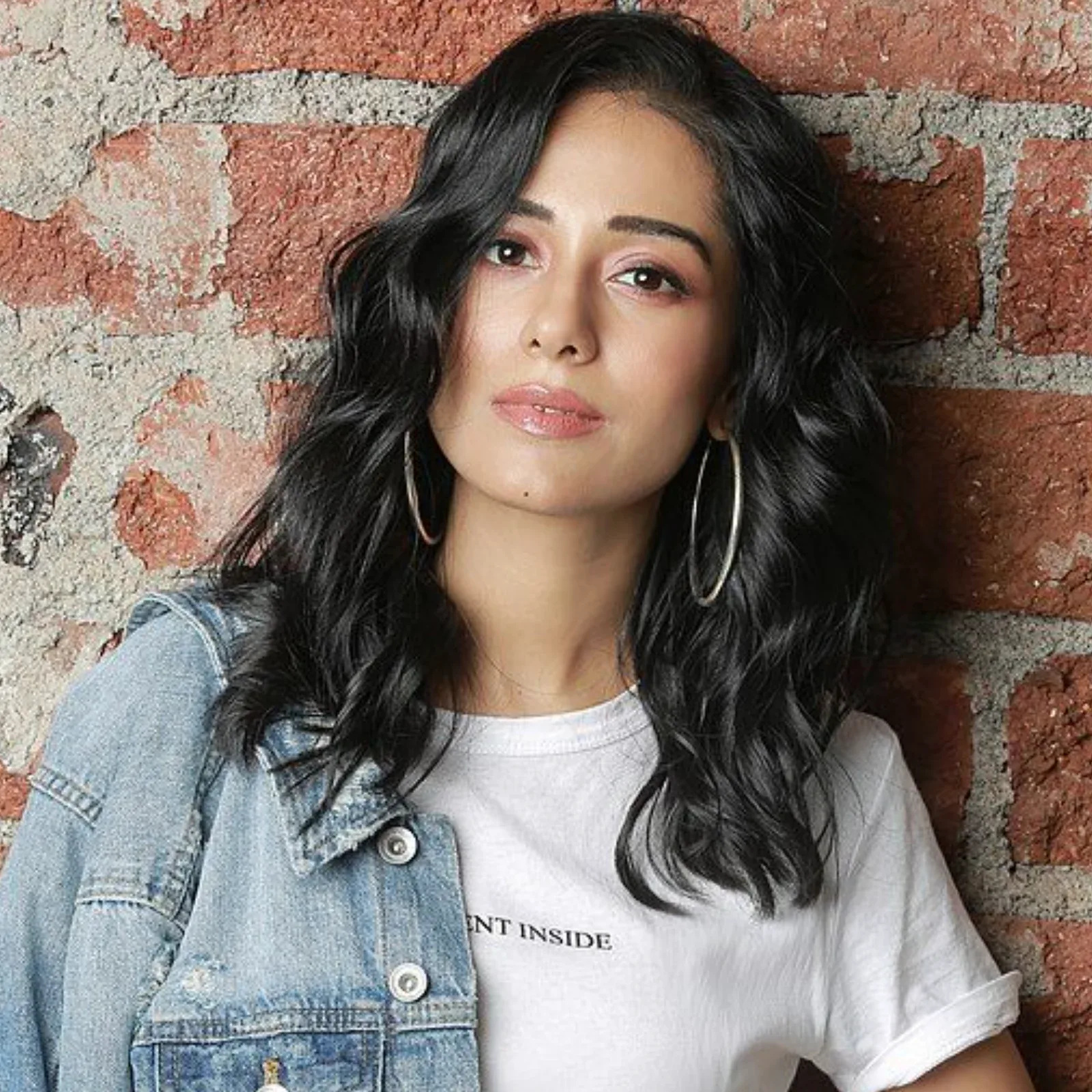 Amrita Rao, an Indian actress, posing against a brick wall, wearing a light blue denim jacket, a white t-shirt, and large silver hoop earrings. She has dark wavy hair and a calm expression.