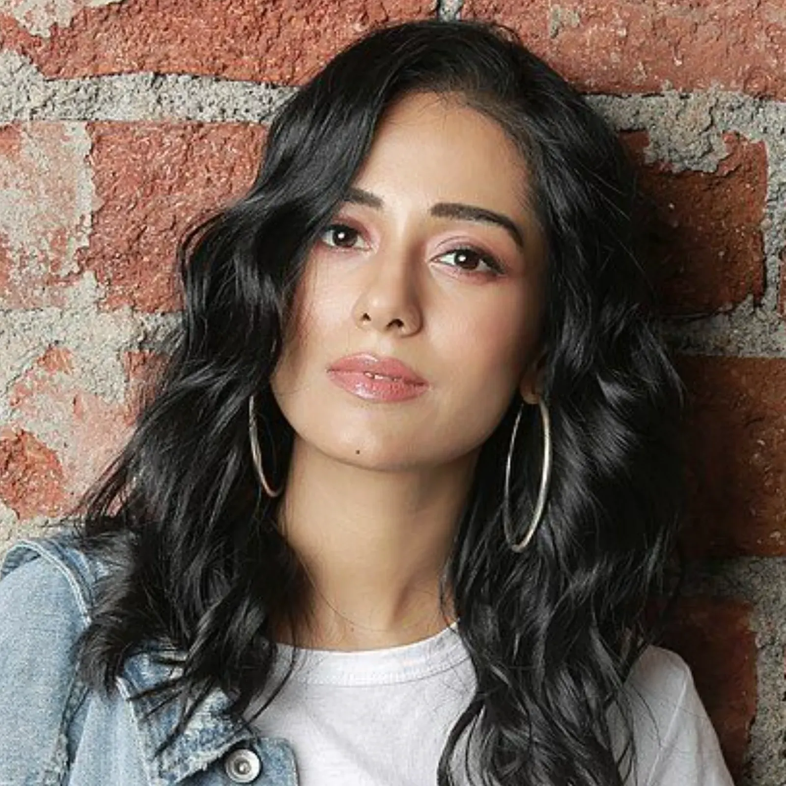 Amrita Rao in a denim jacket and white t-shirt, wearing large silver hoop earrings, posing against a rustic brick wall with a direct gaze.