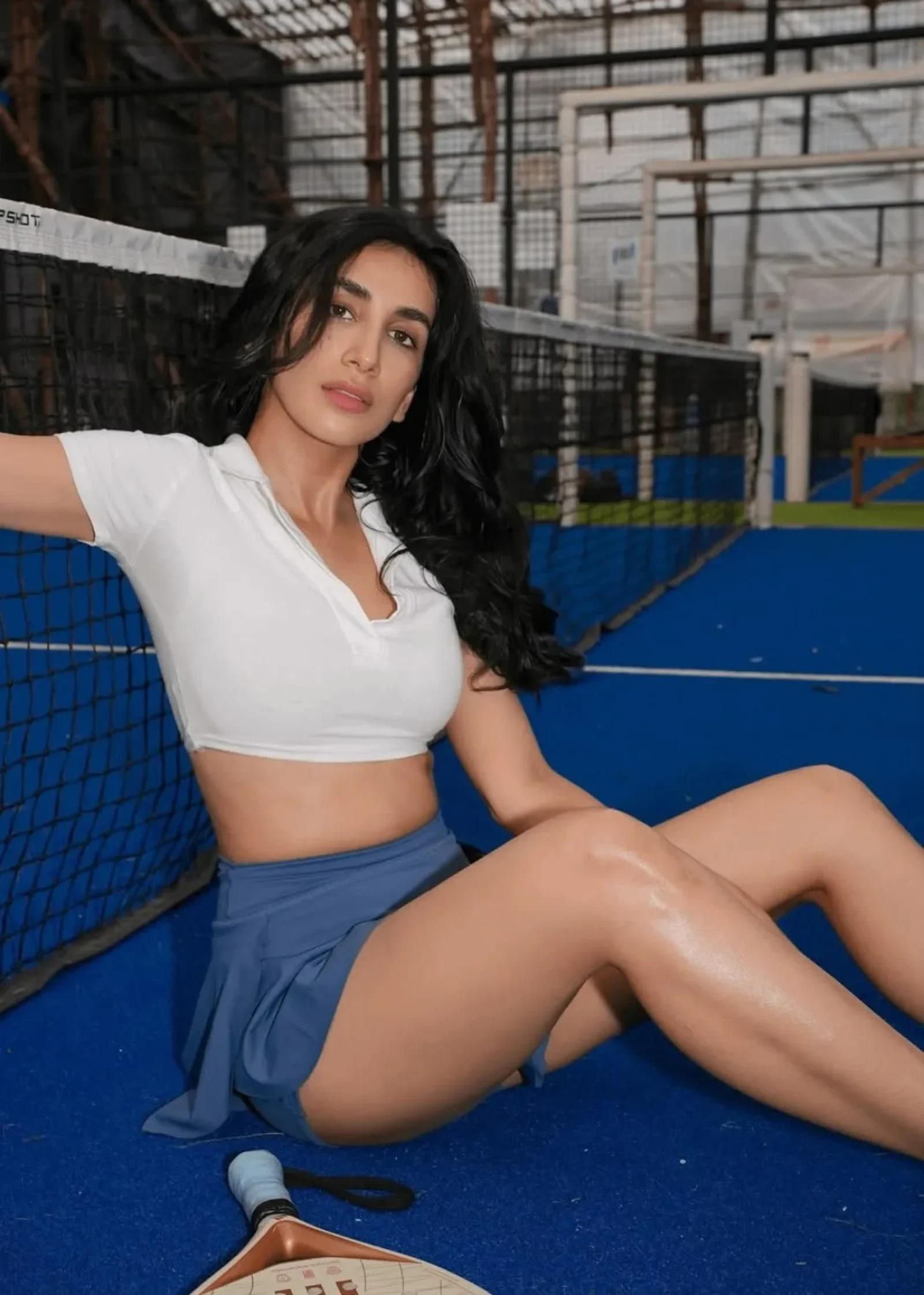 Parul Gulati sitting on a blue padel court, wearing a white cropped polo shirt and a blue pleated mini skirt, looking at the camera.
