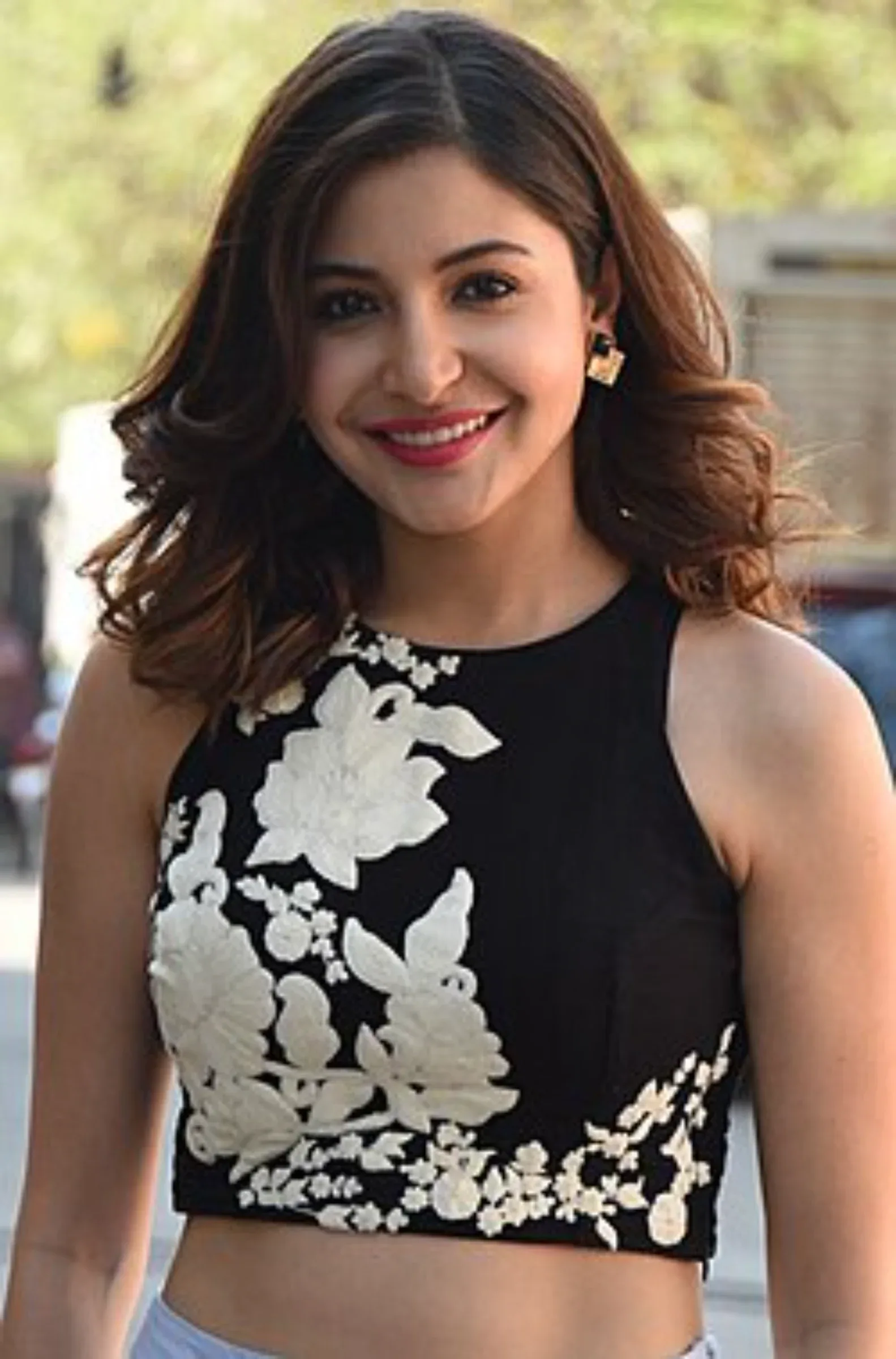 Anushka Sharma smiling brightly, wearing a black sleeveless crop top with white floral embroidery, with her brown wavy hair framing her face.