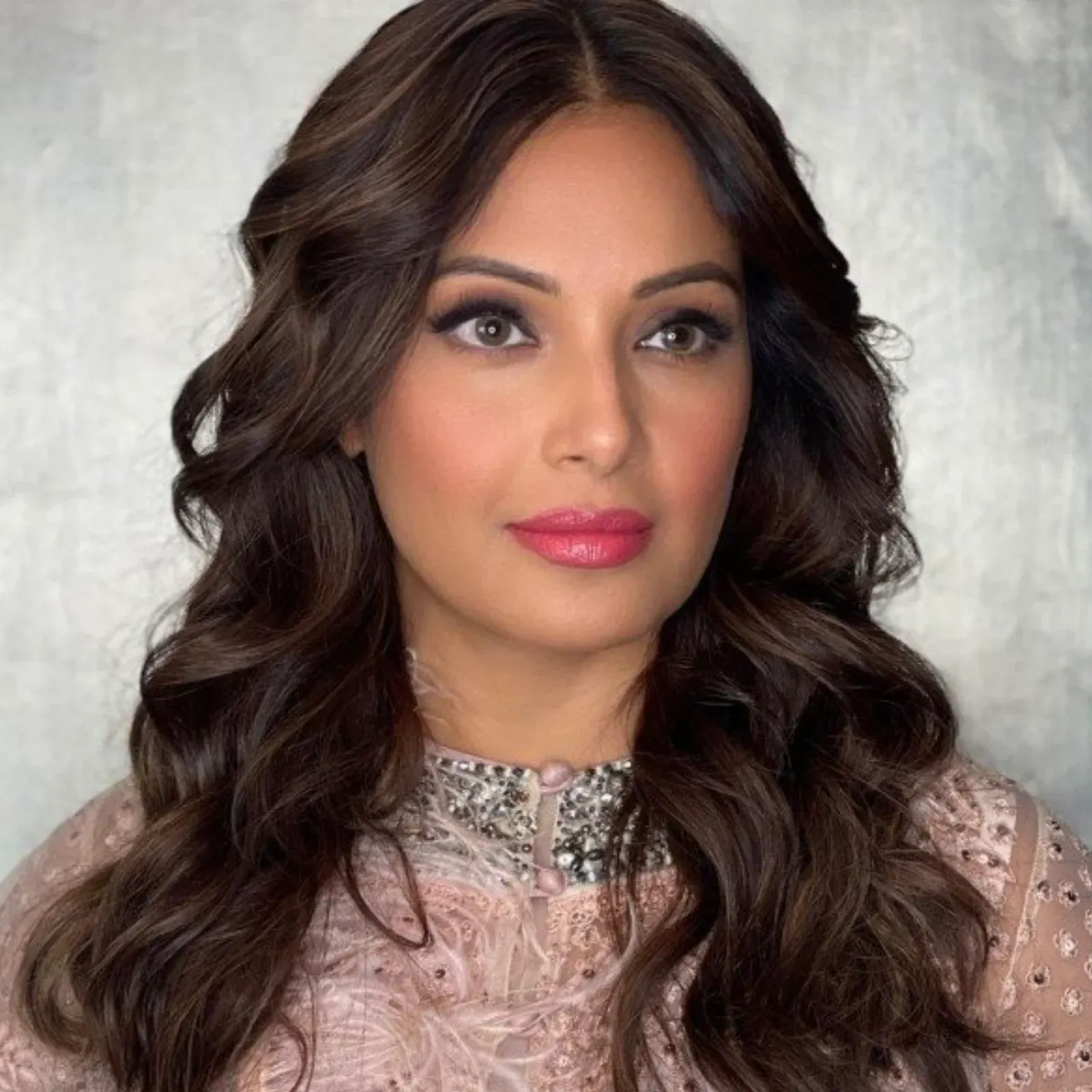 Close-up portrait of Bipasha Basu wearing a light pink embroidered ethnic outfit with a heavily embellished silver and black choker neckline. She has long wavy brown hair, prominent eye makeup, and pink lipstick, looking serene.