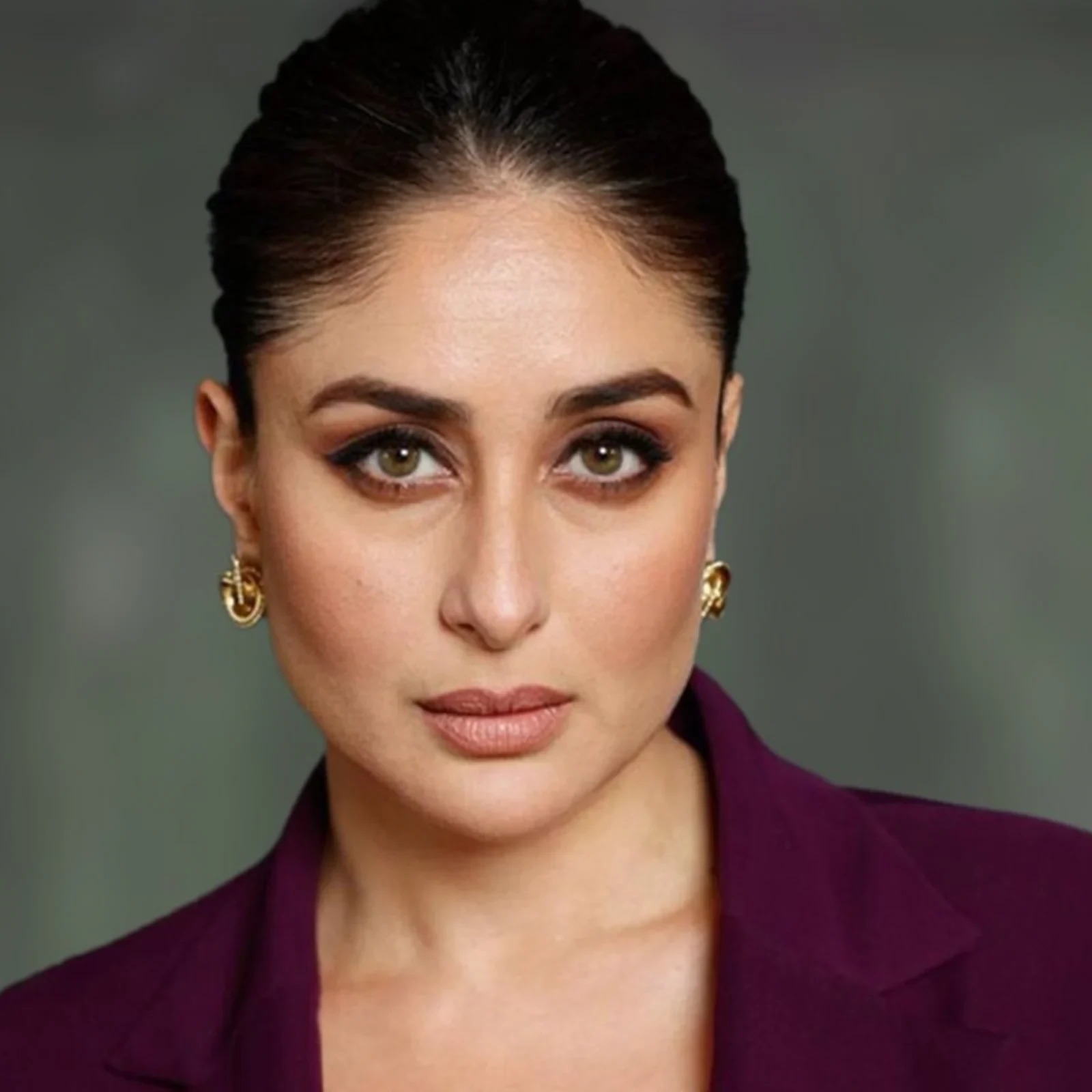 Close-up portrait of Kareena Kapoor wearing a deep purple blazer and gold intricate earrings, with her dark hair pulled back and striking light brown eyes looking directly at the camera.