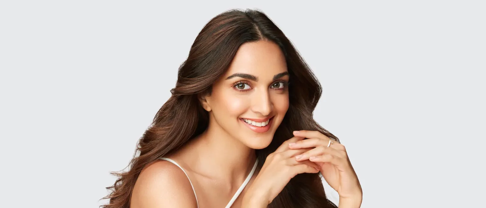 Kiara Advani smiling brightly, looking directly at the camera, with long wavy dark brown hair and wearing a white top. Her hands are clasped gently.